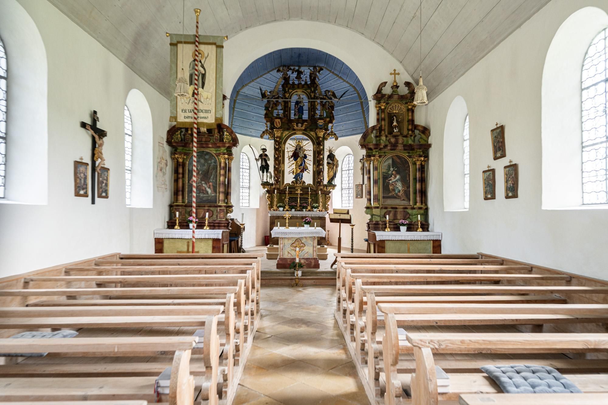 Innenansicht einer hellen Kapelle mit Holzbänken und drei reich verzierten Altären. An den weißen Wänden hängen Bilder und ein Kruzifix.