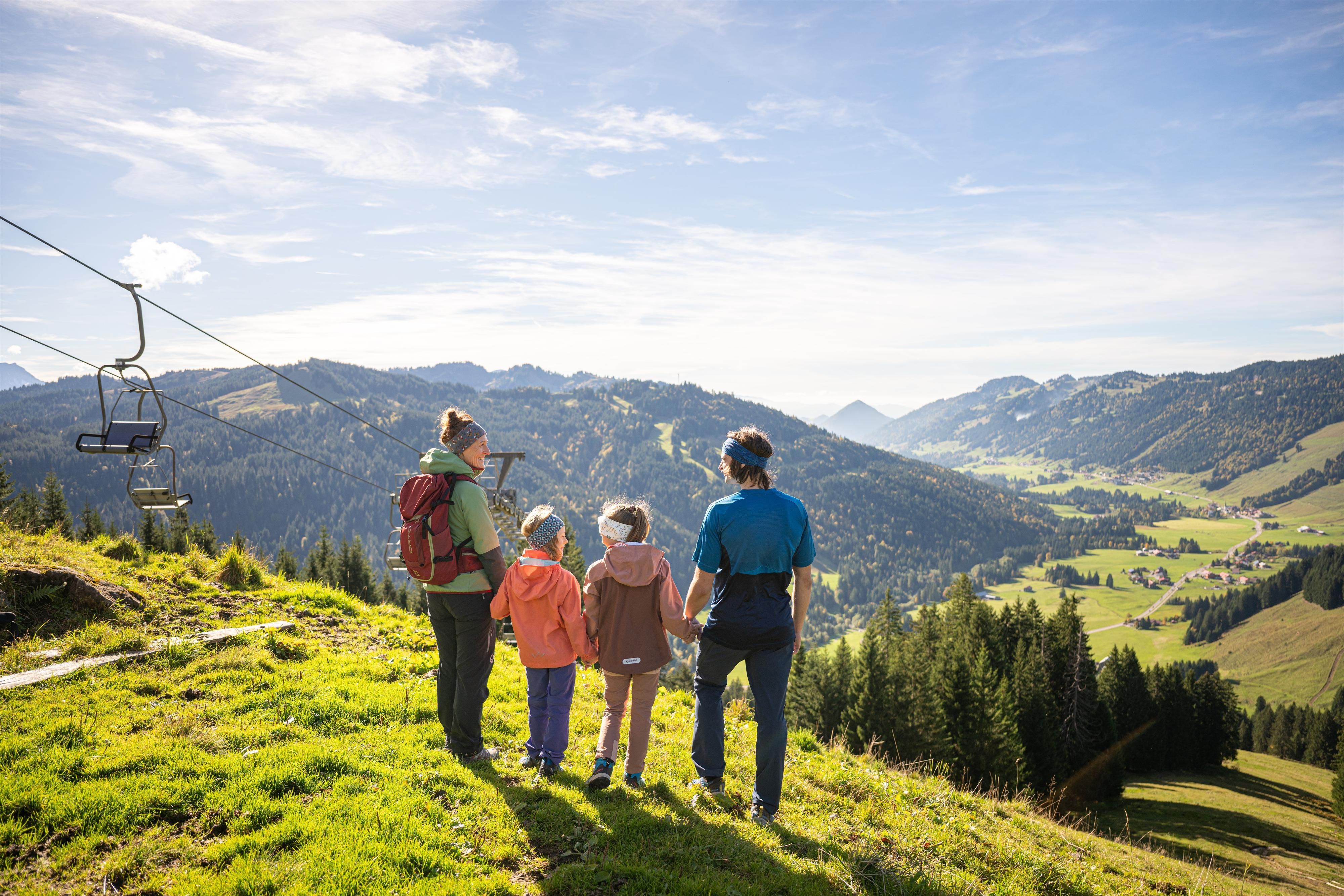 Eine Familie mit zwei Kindern steht auf einer sonnigen Bergwiese und blickt gemeinsam über ein weites Tal mit Wäldern, Hügeln und kleinen Dörfern. Links im Bild verläuft ein Sessellift den Hang hinauf, darüber ist ein heller blauer Himmel mit dünnen Wolken zu sehen.