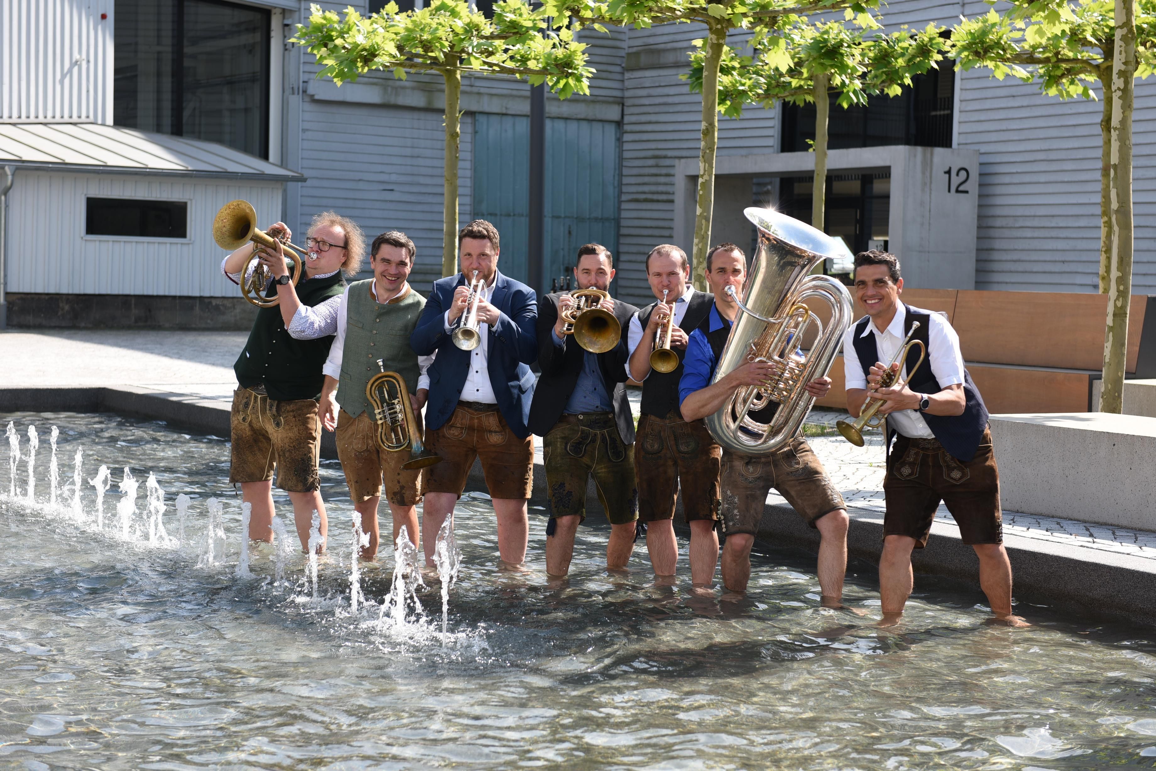 Die 7köpfige Band steht in einem Brunnen mit wenig Wasser, alle tragen bayerische Lederhosen und Ihre Instrumente.