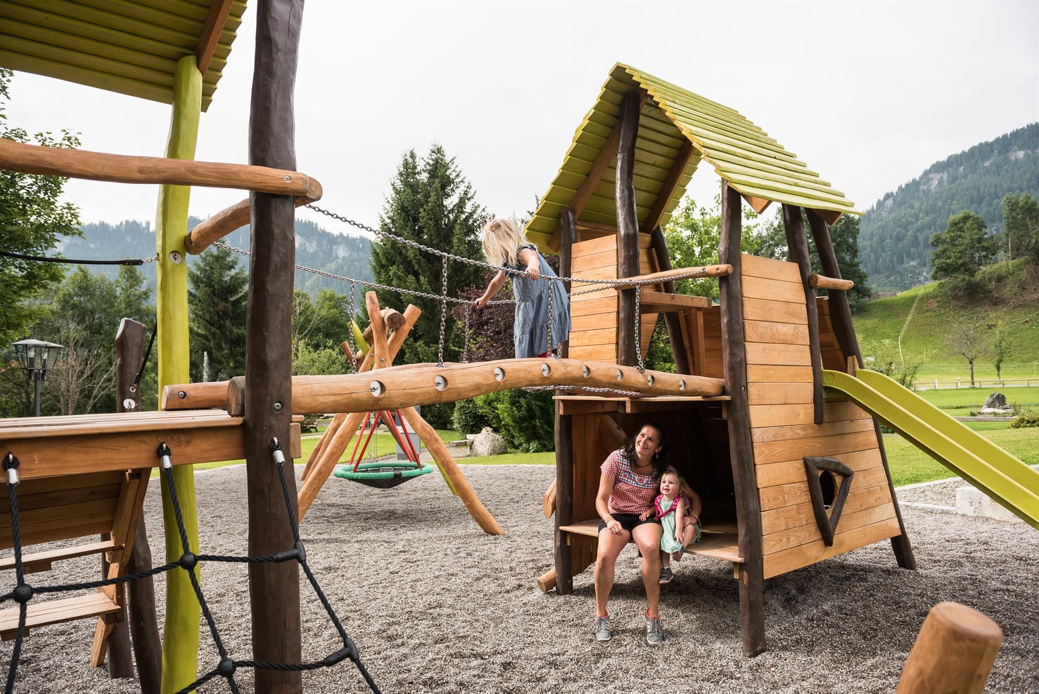 Holzspielplatz mit Kletterturm, Hängebrücke, Rutsche und Schaukel, eine Frau und zwei Kinder interagieren.