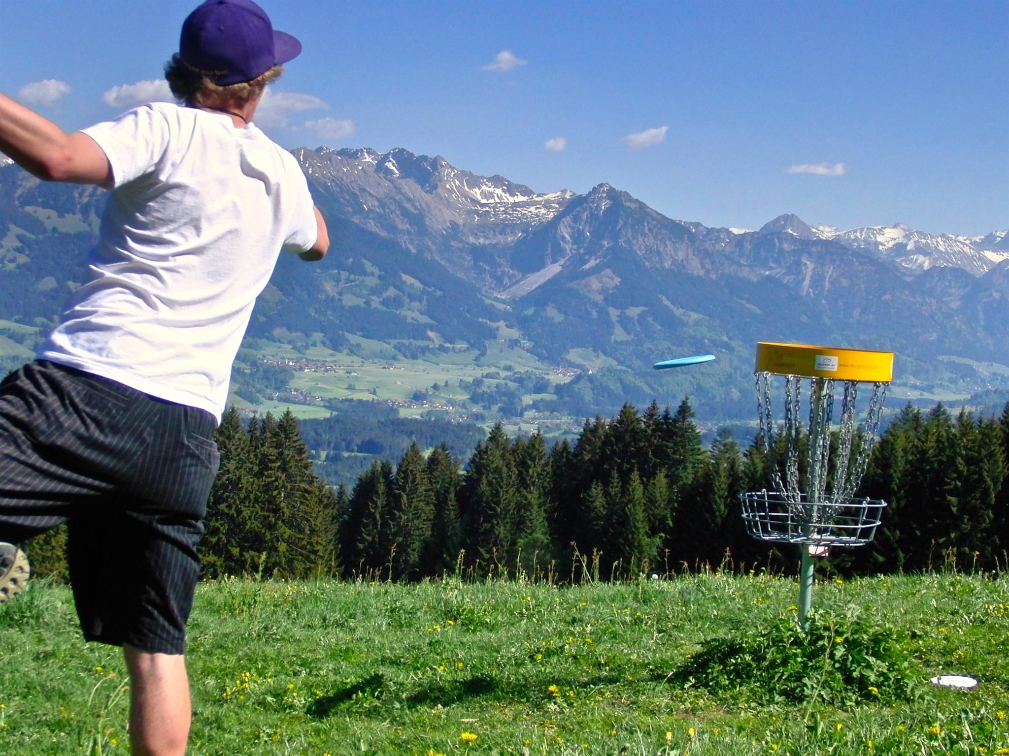Mann wirft blaue Frisbeescheibe auf gelben Disc Golf Korb.