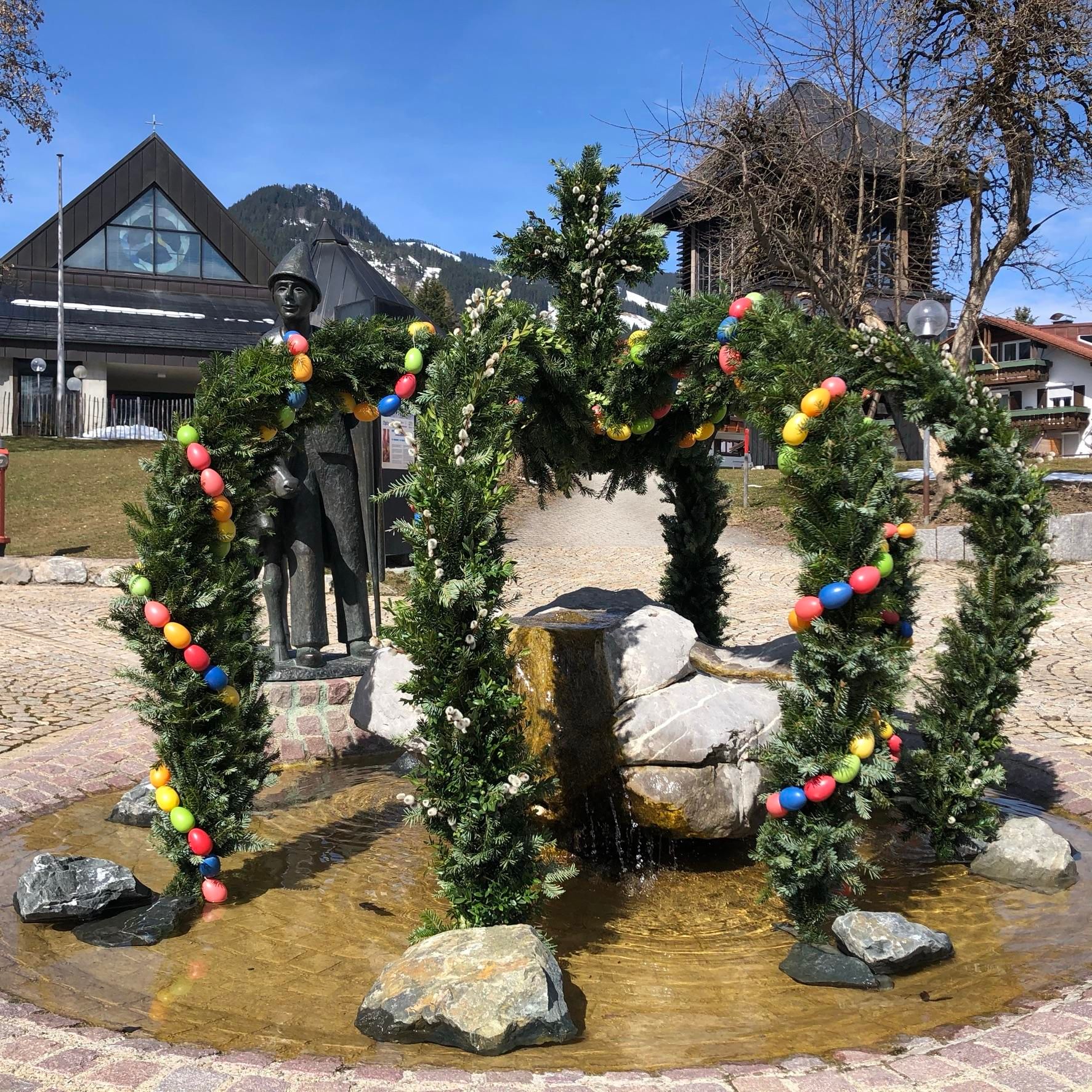 Osterbrunnen, geschmüclt mit kleinen Tannenbäumen und bunten Ostereiern. vor Pfarrkirche St. Urlich in Obermaiselstein.
