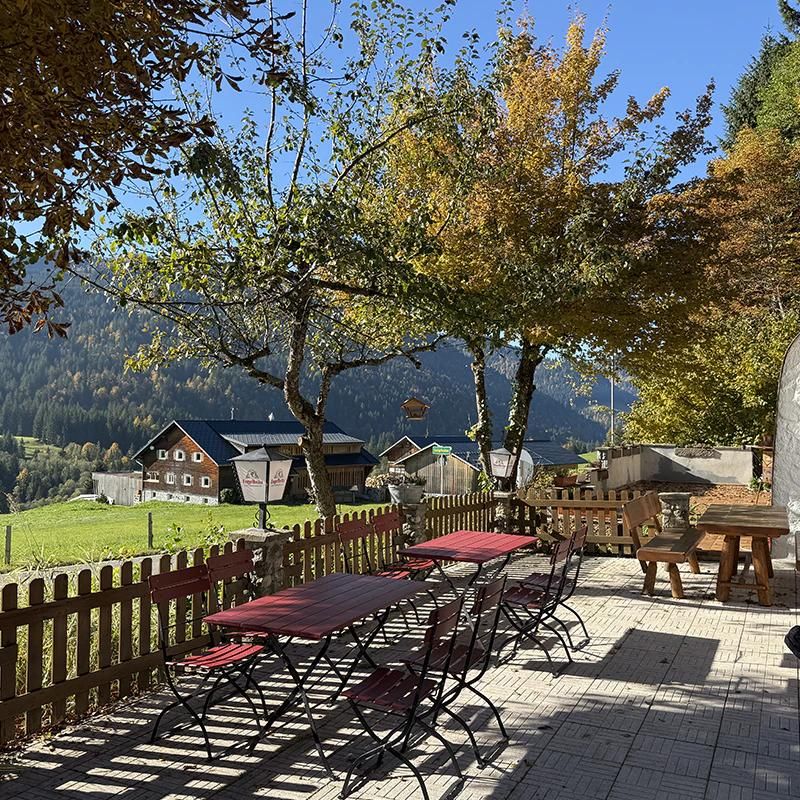 Eine gemütliche Terrasse mit Tischen und Stühlen, umgeben von buntem Laub. Im Hintergrund sieht man grüne Wiesen und Berge.