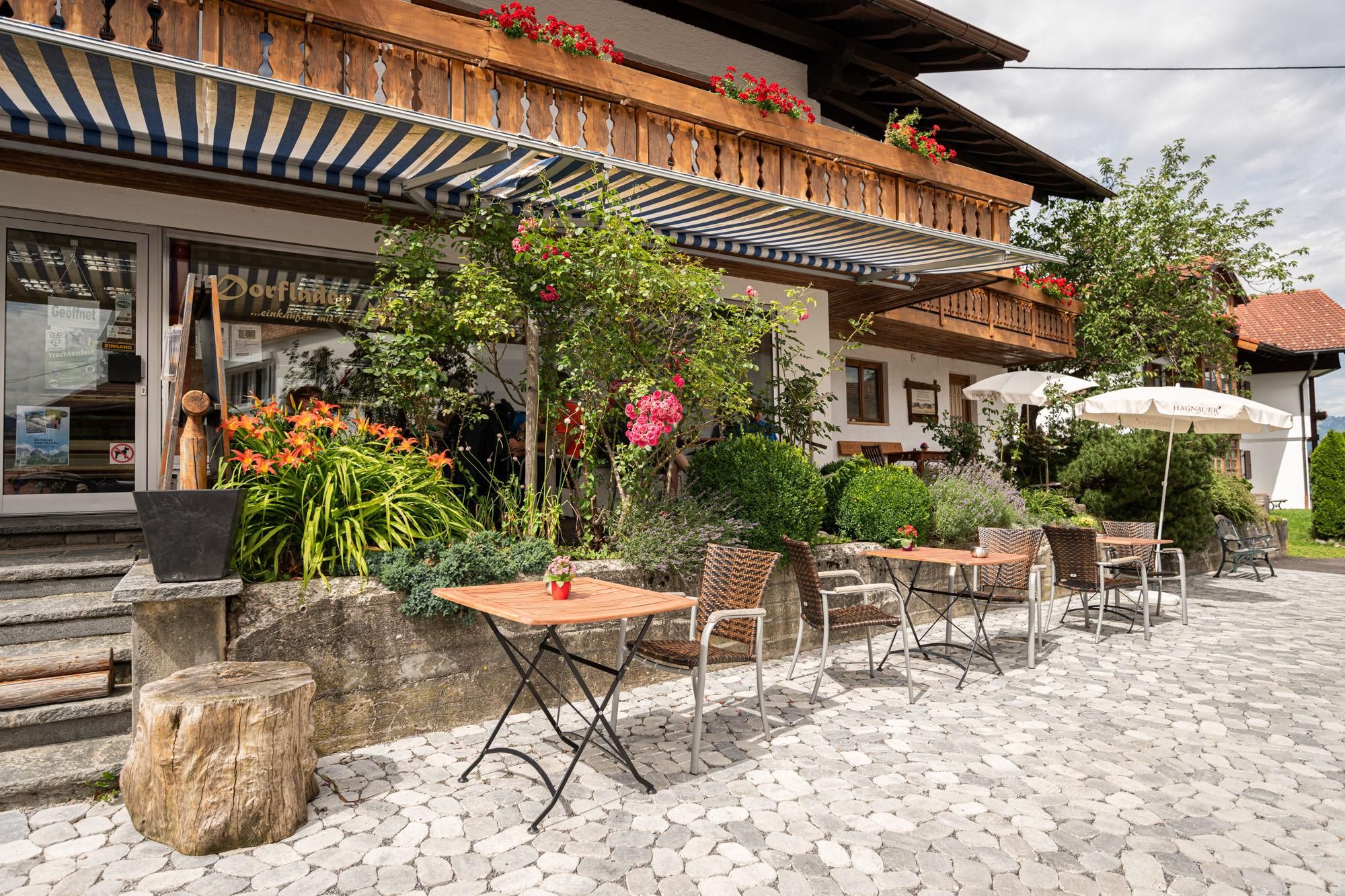 Ein gemütlicher Außenbereich am Dorfladen in Bolsterlang mit Tischen und Stühlen. Üppige Pflanzen und Blumen sowie ein Sonnenschirm verleihen der Atmosphäre einen einladenden Charme.