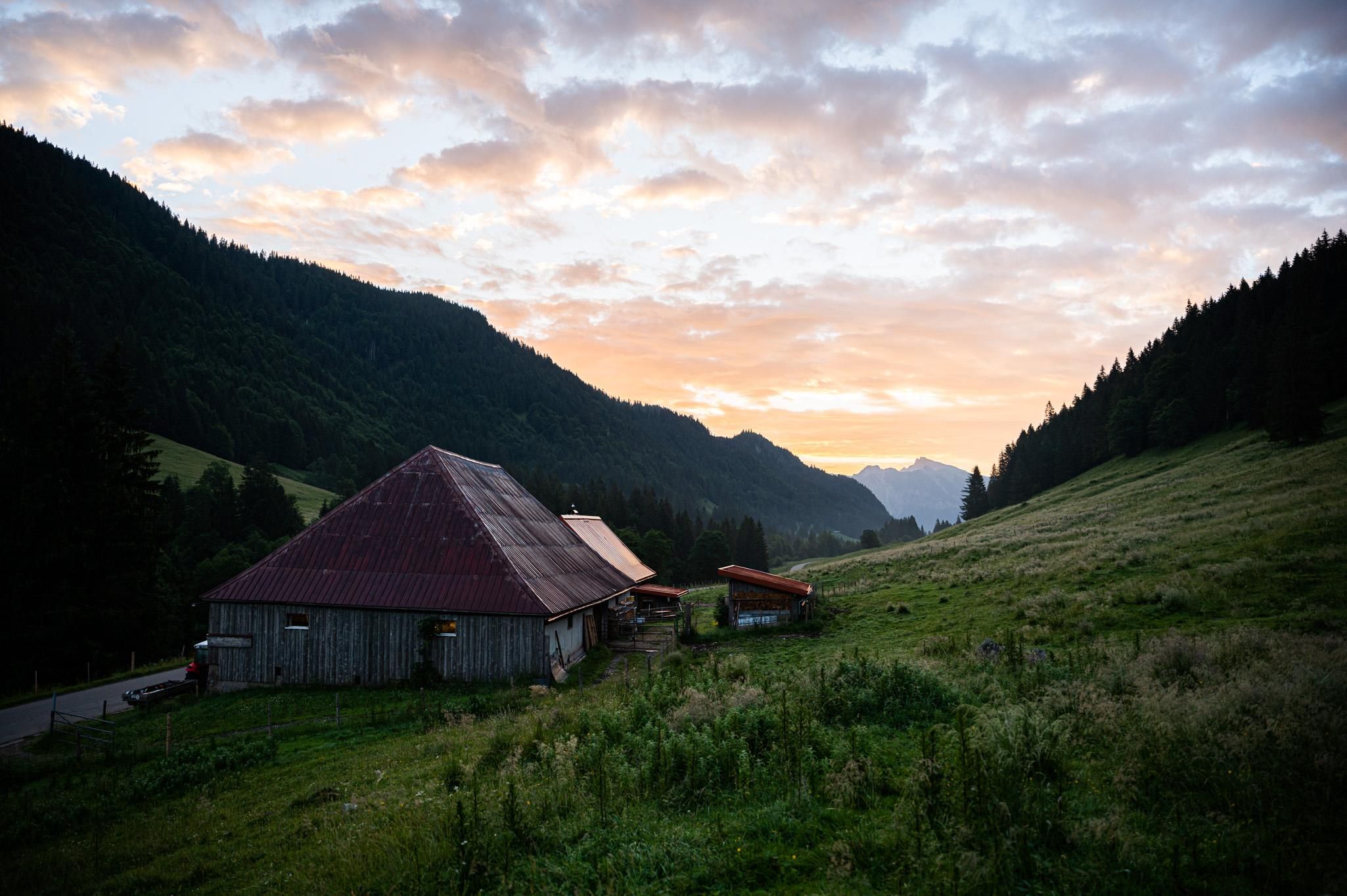 Hölzerne Sennalpe mit rotem Dach auf grüner Wiese, umgeben von bewaldeten Bergen, Abenddämmerung mit bunten Wolken.