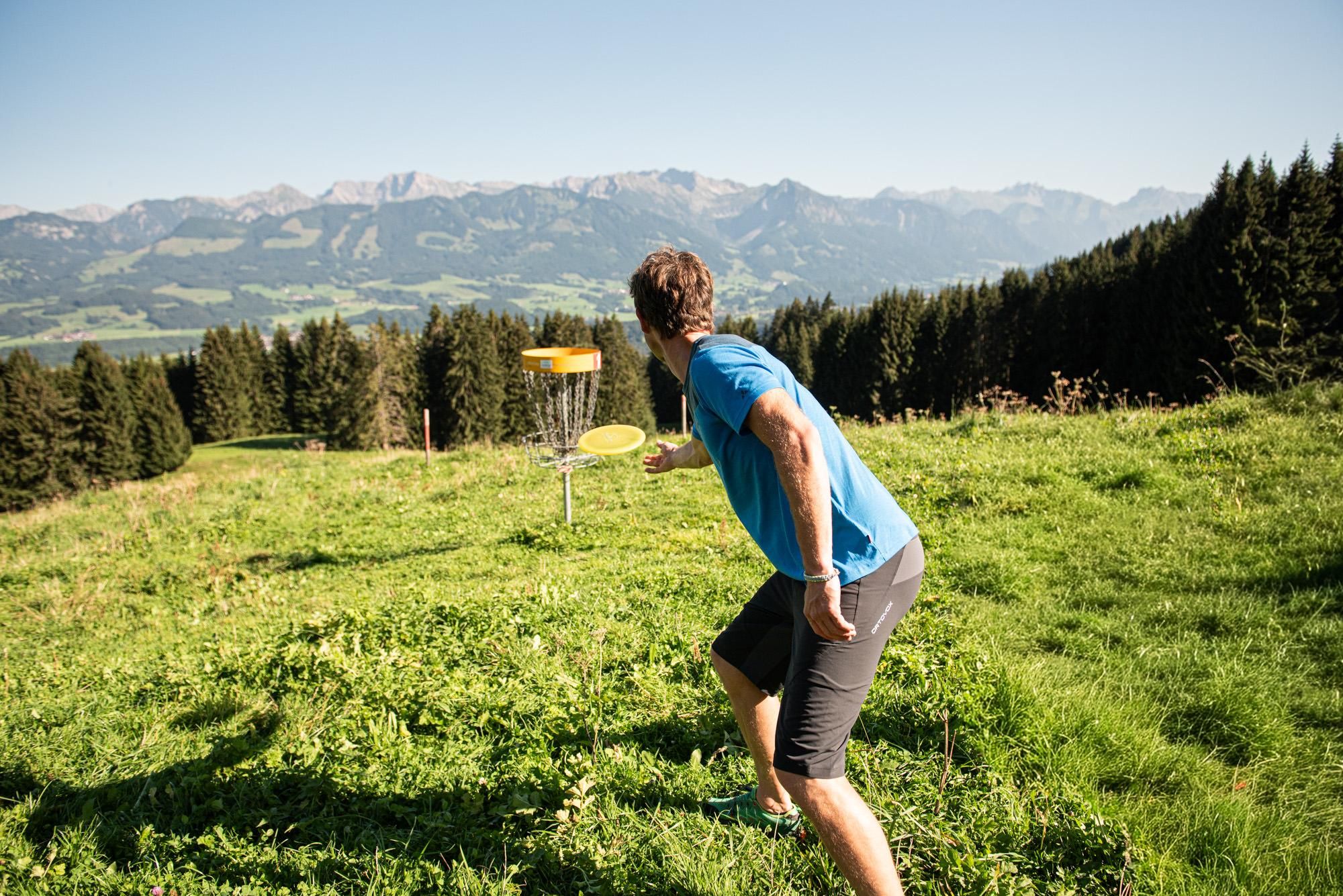 Ein Mann spielt Disc Golf auf einer Wiese in den Bergen. Im Hintergrund sind die Berge und ein klarer Himmel zu sehen.