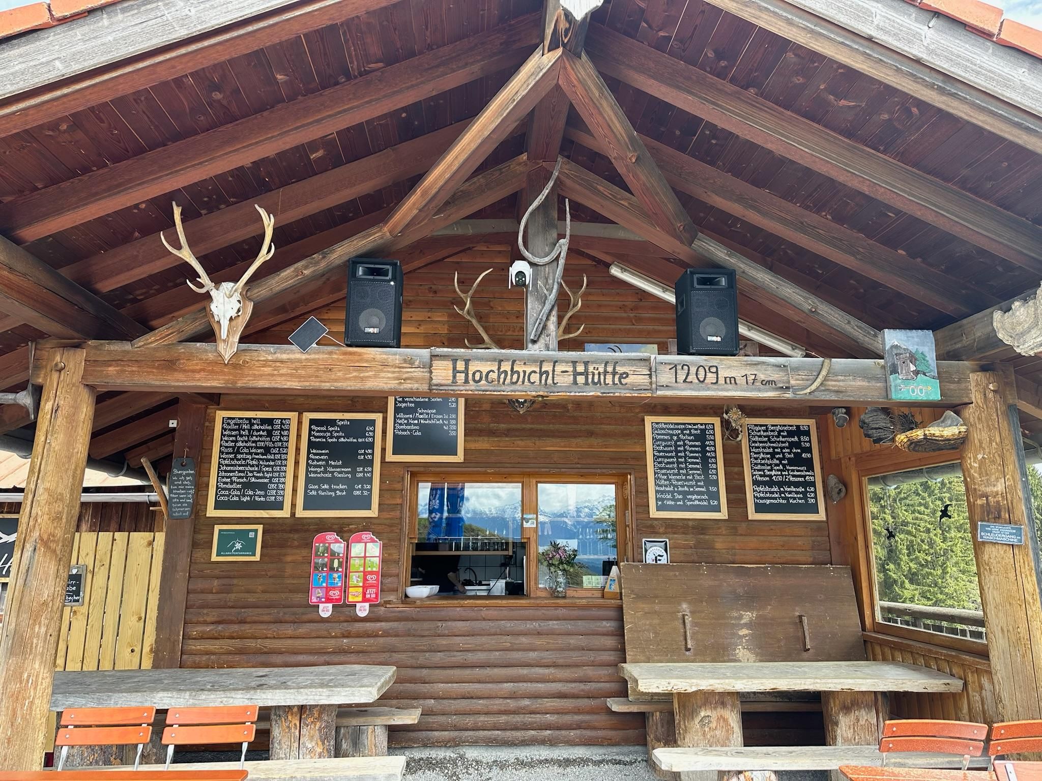 Eine rustikale Berghütte mit Holzverkleidung und einem Schild. Auf der Terasse stehen Tische und es sind einige schilfrohrartige Dekorationen und Beschilderungen sichtbar.