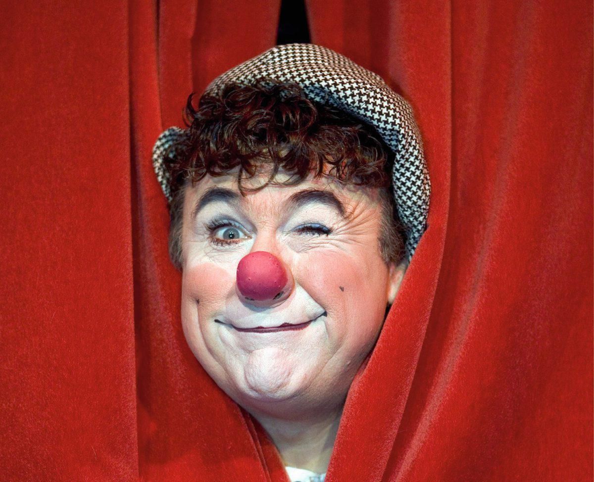Ein fröhlicher Clown mit roter Nase und karierter Mütze lugt durch einen roten Vorhang und zwinkert mit einem Auge.