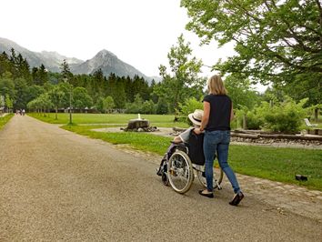 Frau schiebt Rollstuhl durch den Kurpark Fischen in den Hörnerdörfern im Allgäu. Hinten Berge, rechts Barfußpfad. 