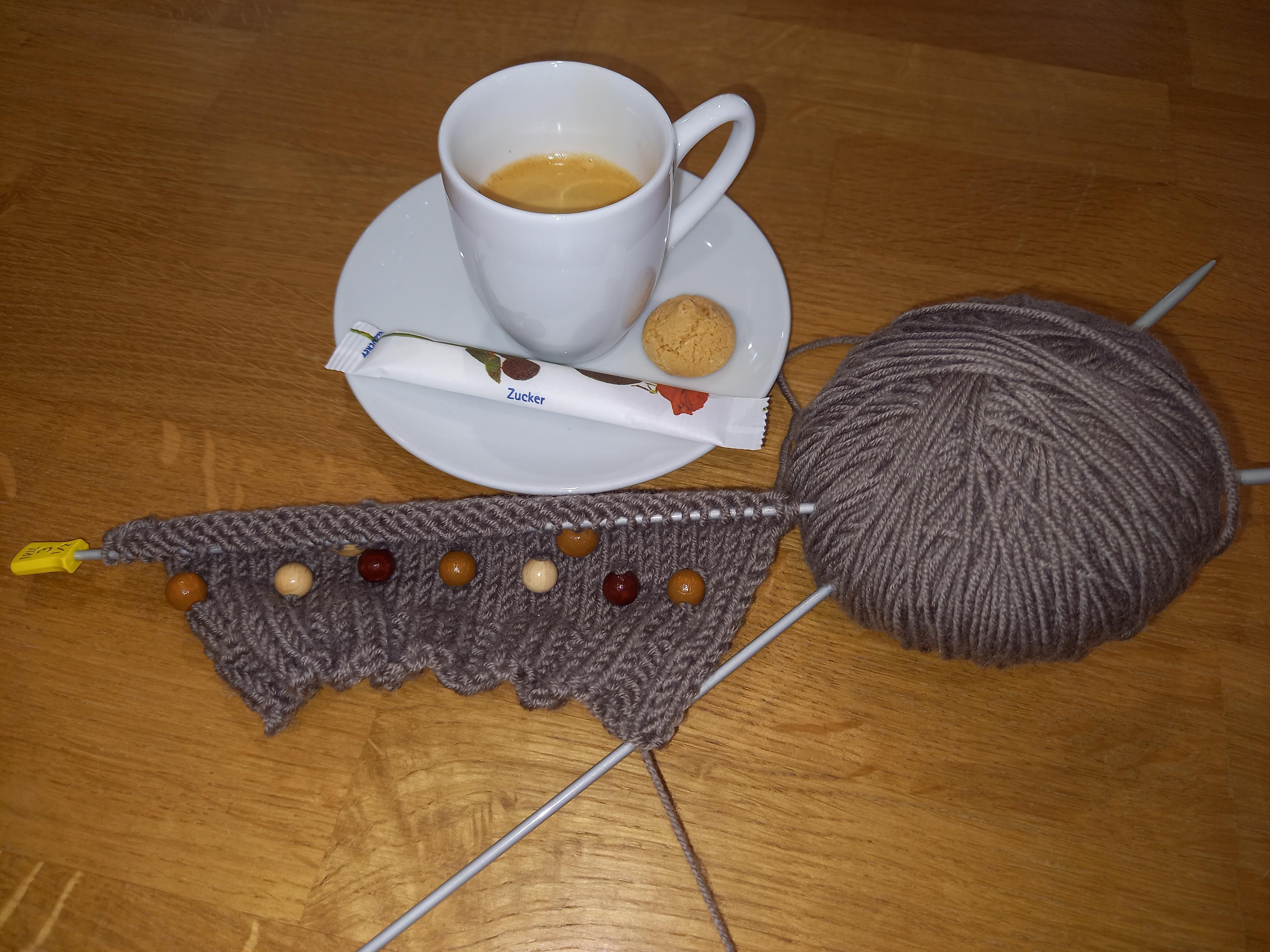 Eine Tasse Espresso, auf dem Unterteller ein Zuckerpäckchen und ein Mandelkeks, daneben Strickzeug mit grauer Wolle.