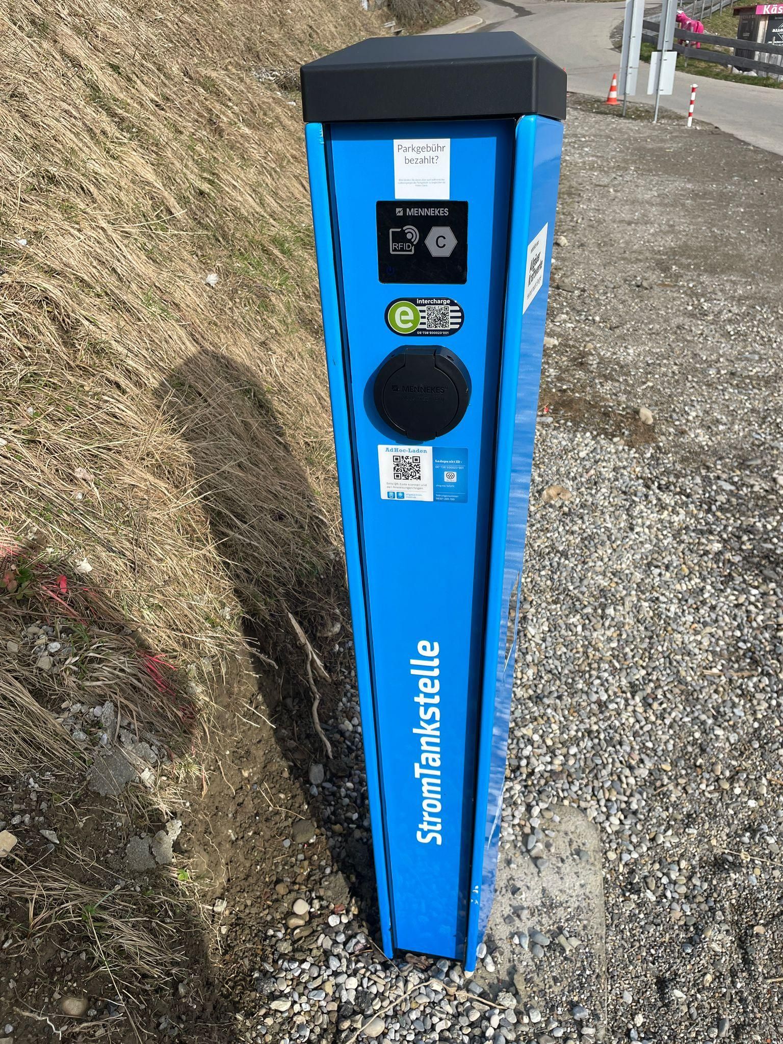 Eine blaue Stromtankstelle mit schwarzem Kopf steht auf einem Schotteruntergrund neben einem grasbewachsenen Hang. Verschiedene Symbole und Schriftzüge sind auf der Säule angebracht.