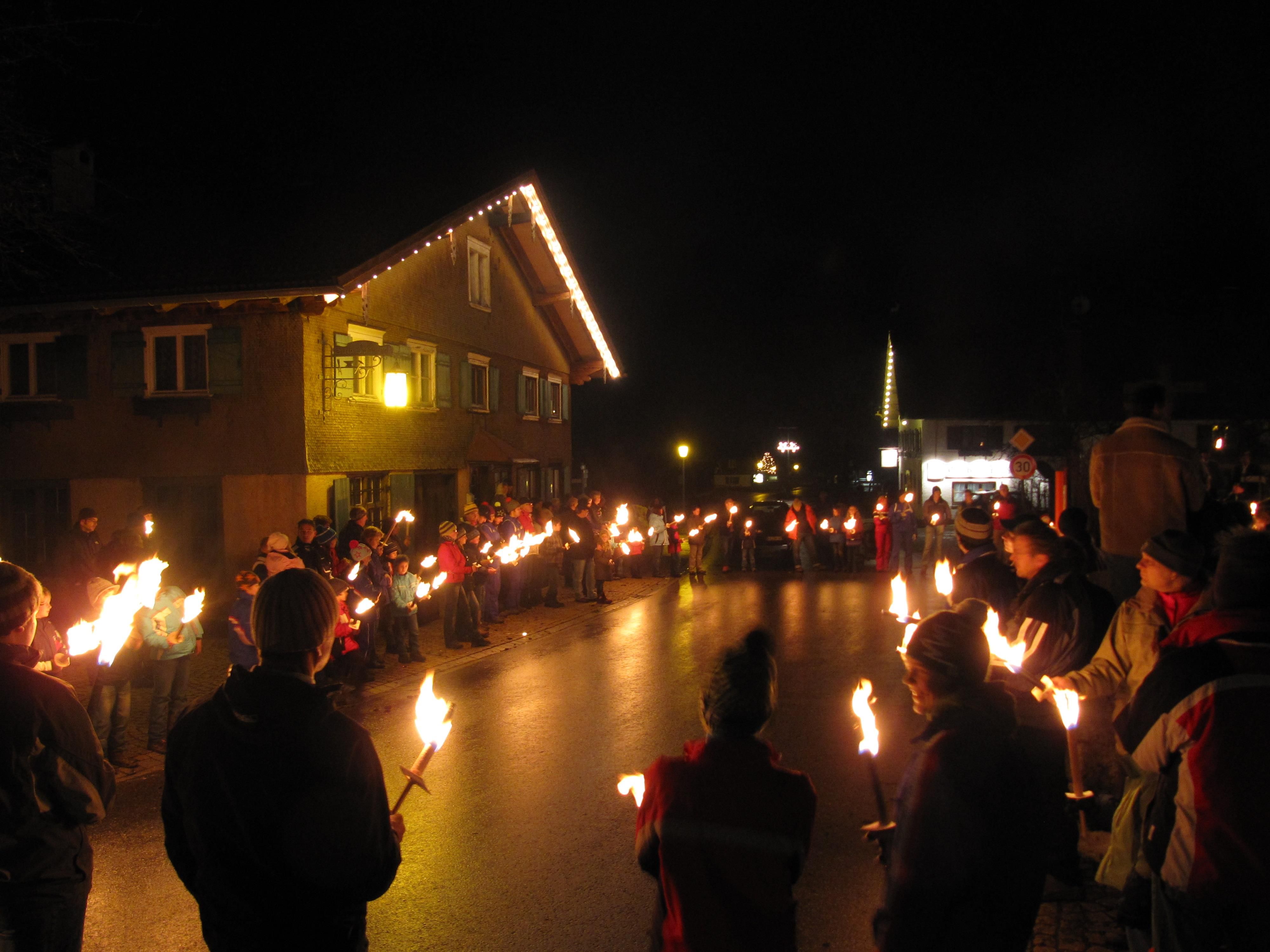 Menschen versammeln sich nachts auf einer Straße zum traditionellen Fackellauf. Häuser sind beleuchtet, der Himmel ist dunkel.