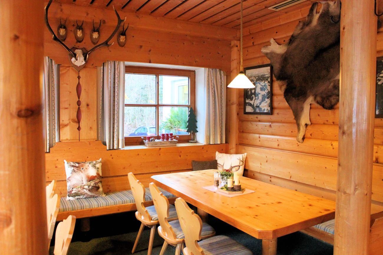 Rustikale Gaststube. Holzwände und -decke. Heller Holztisch mit Stühlen und Eckbank. Dekoriert mit Hirschgeweih und Tierfell. Fenster mit Vorhängen.