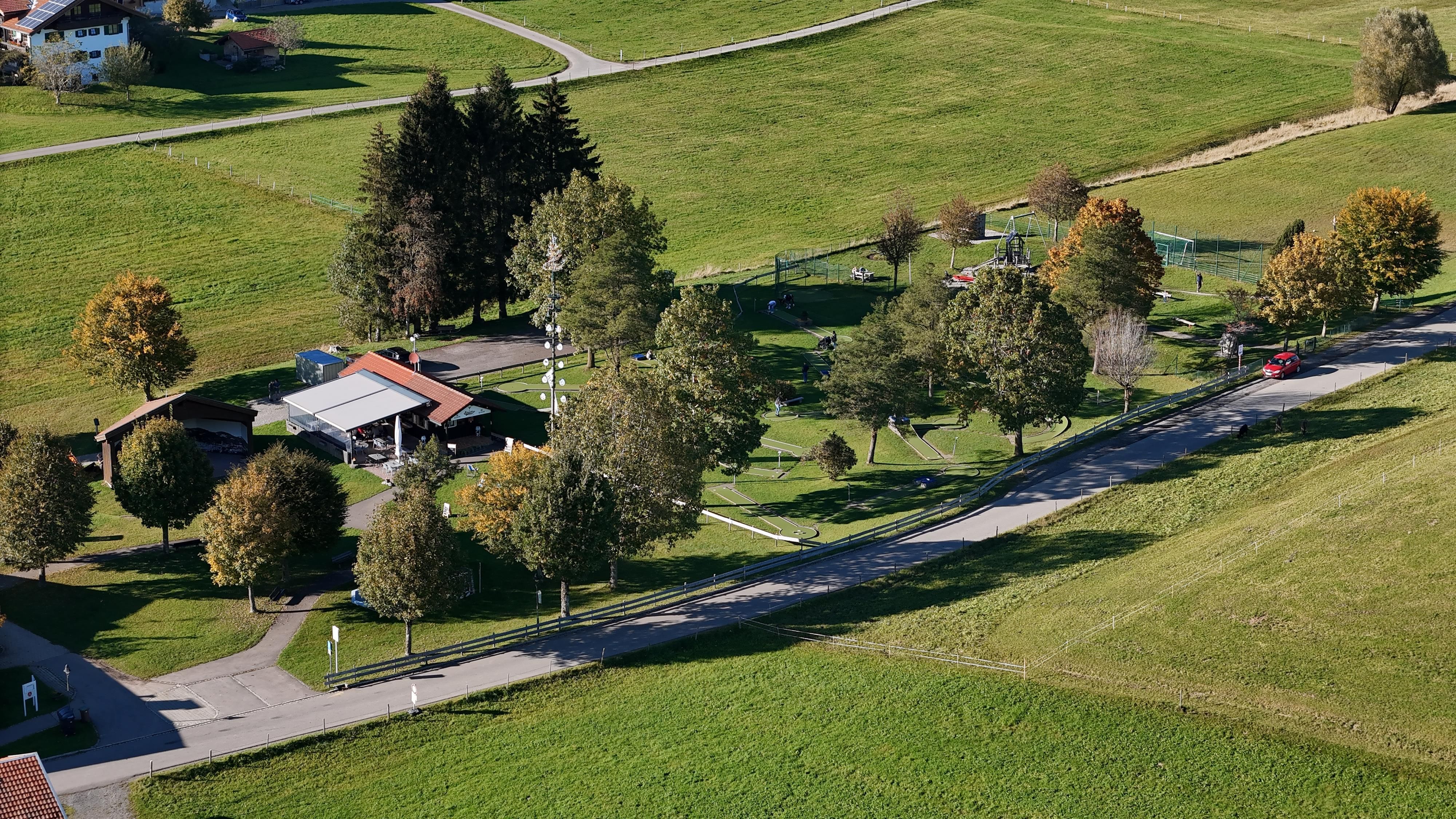 Luftaufnahme von Schickie's Minigolfanlage, eingebettet in eine grüne Landschaft mit Minigolfbahnen, einem Spielplatz mit bunten Geräten, umgeben von Bäumen und Wegen.