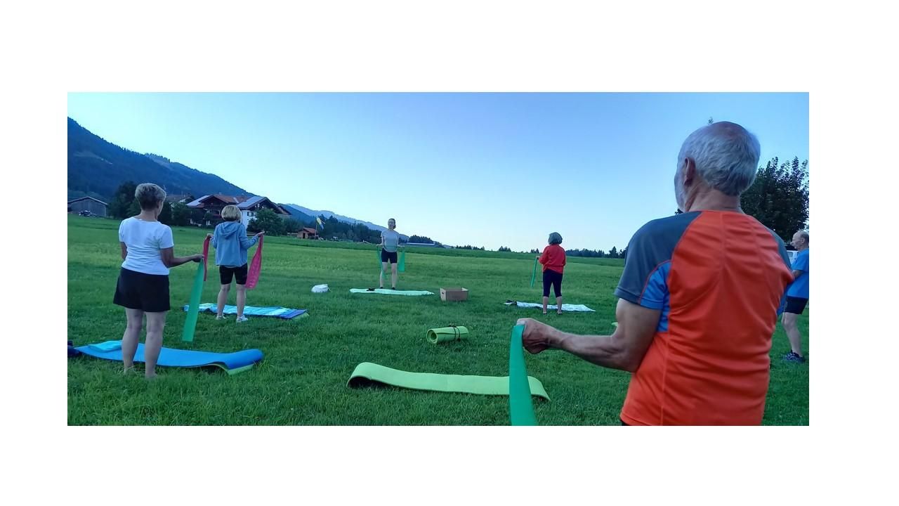 Mehrere Personen machen Gymnastik im Freien auf einer grünen Wiese mit Bergen im Hintergrund.
