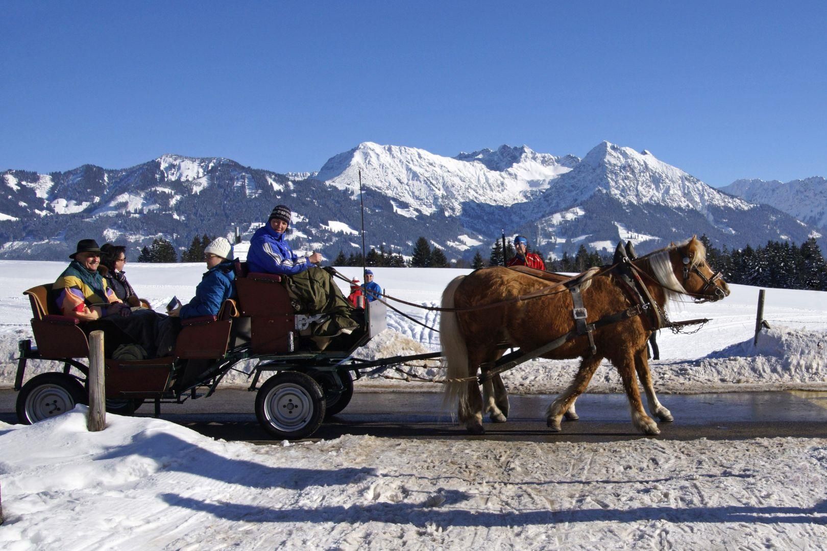 Eine Pferdekutsche mit Passagieren fährt auf einer schneebedeckten Straße, im Hintergrund schneebedeckte Berge.