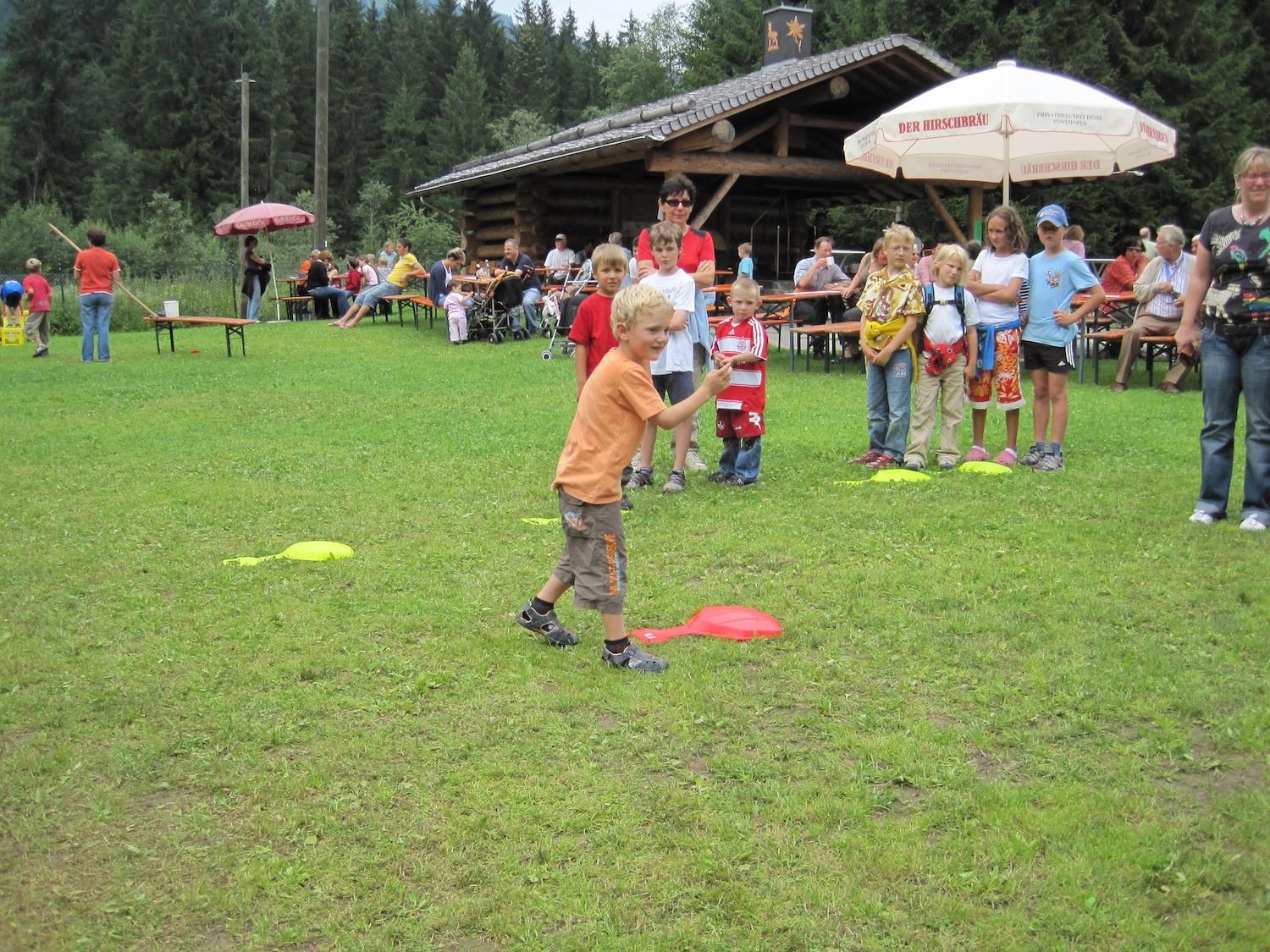 Familienfest auf einer grünen Wiese. Ein Junge in orangefarbenem T-Shirt spielt mit einem roten Gegenstand. Im Hintergrund stehen Kinder und Erwachsene.