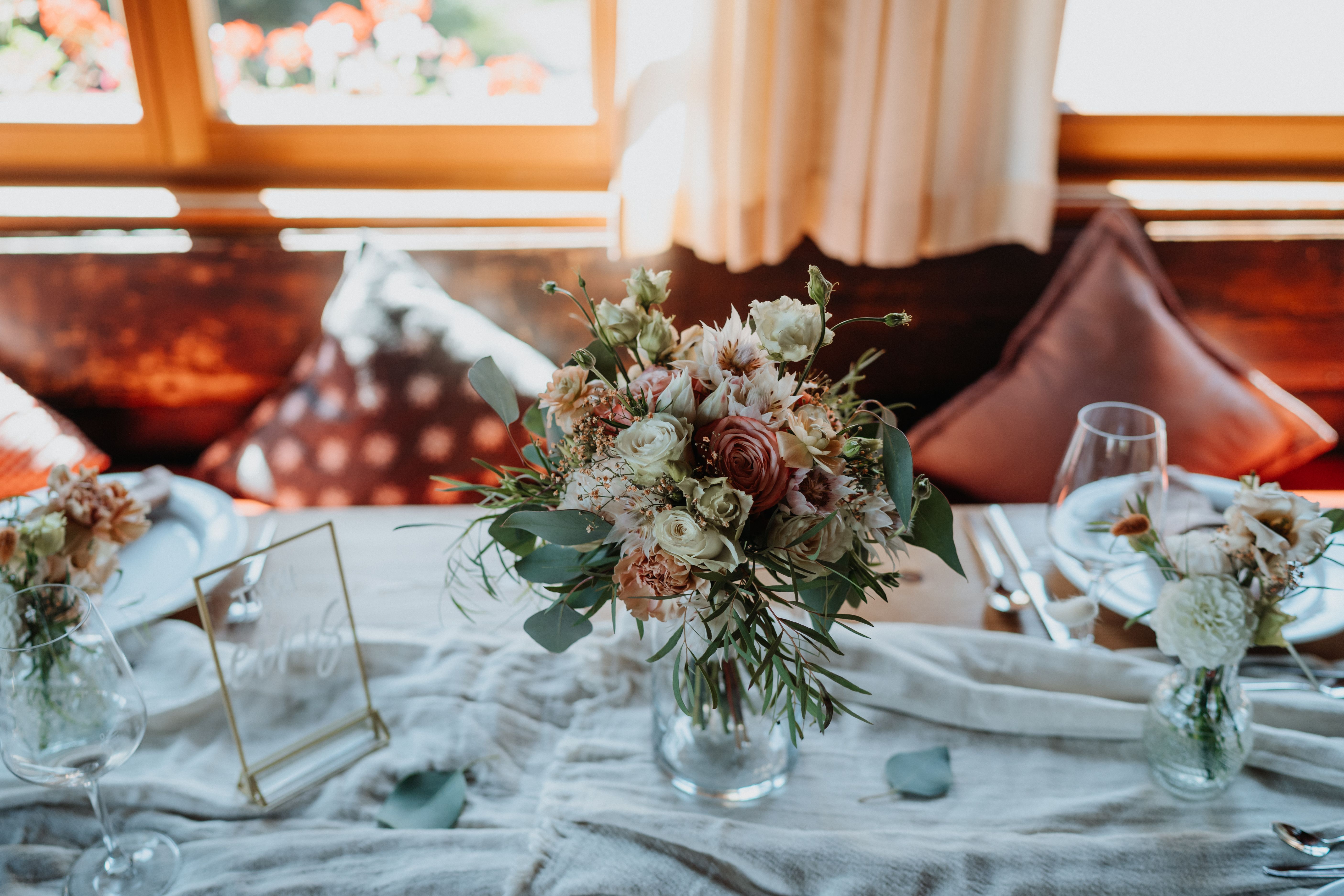Blumengesteck auf eingedecktem Tisch, dahinter Sitzbank und Fenster.