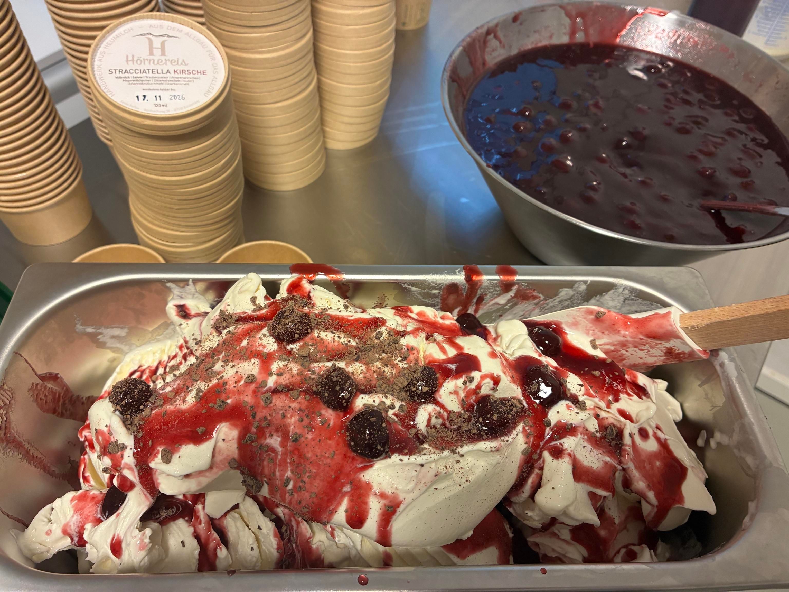 Ein Behälter mit cremigem Eis, garniert mit roter Fruchtsauce und Beeren. Im Hintergrund stehen leere Eisbecher und eine Schüssel mit weiterer Sauce.