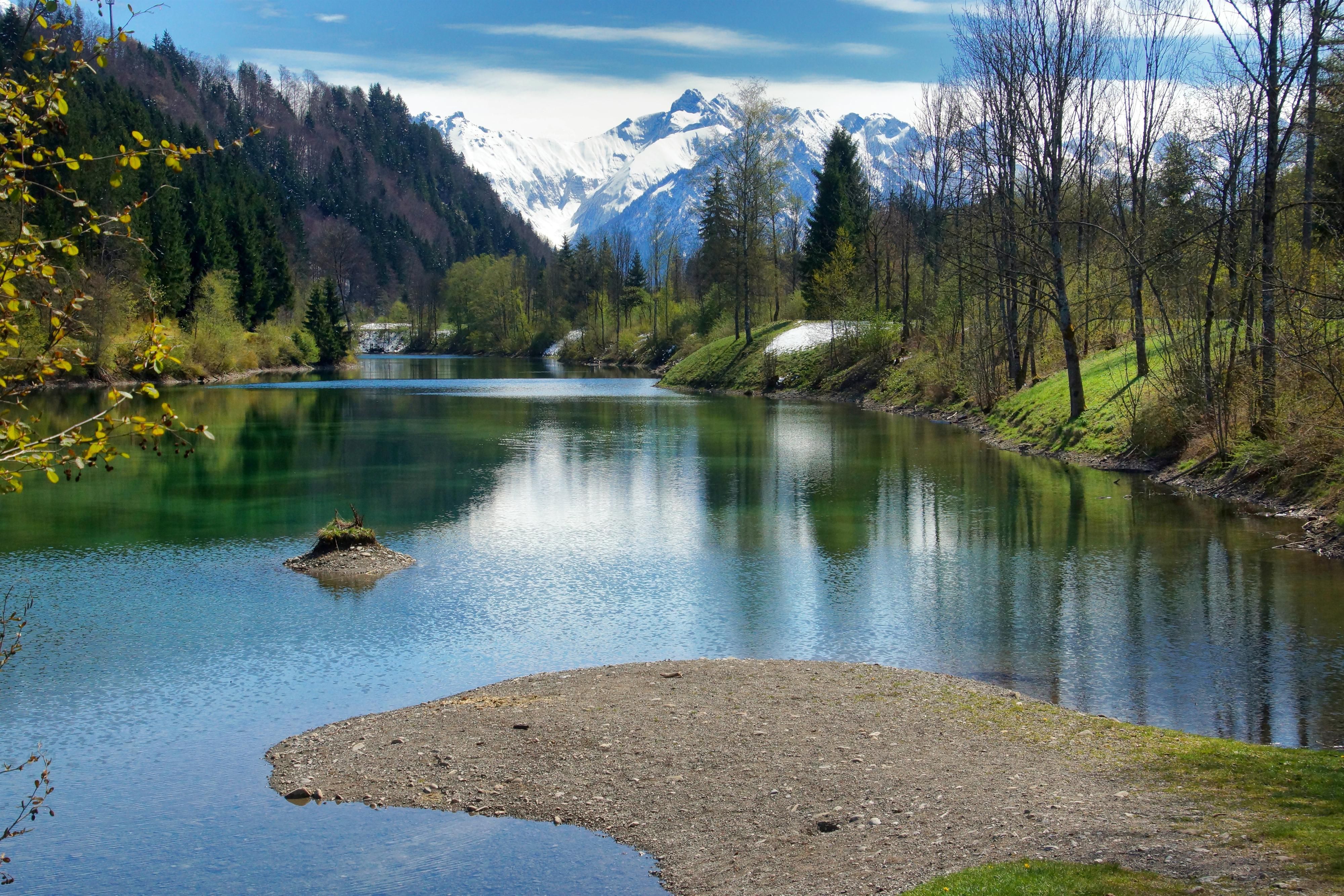 Auwaldsee im Frühling, das Wasser steht relativ niedrig, die Bäume haben noch keine Blätter, die Berge sind verschneit.