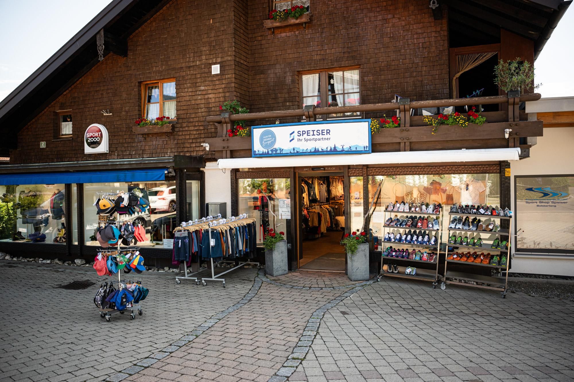 Ein Geschäft mit einer Holzfassade, das Outdoor-Ausrüstung verkauft. Vor dem Laden sind Kleiderständer und Regale mit Schuhen aufgestellt.