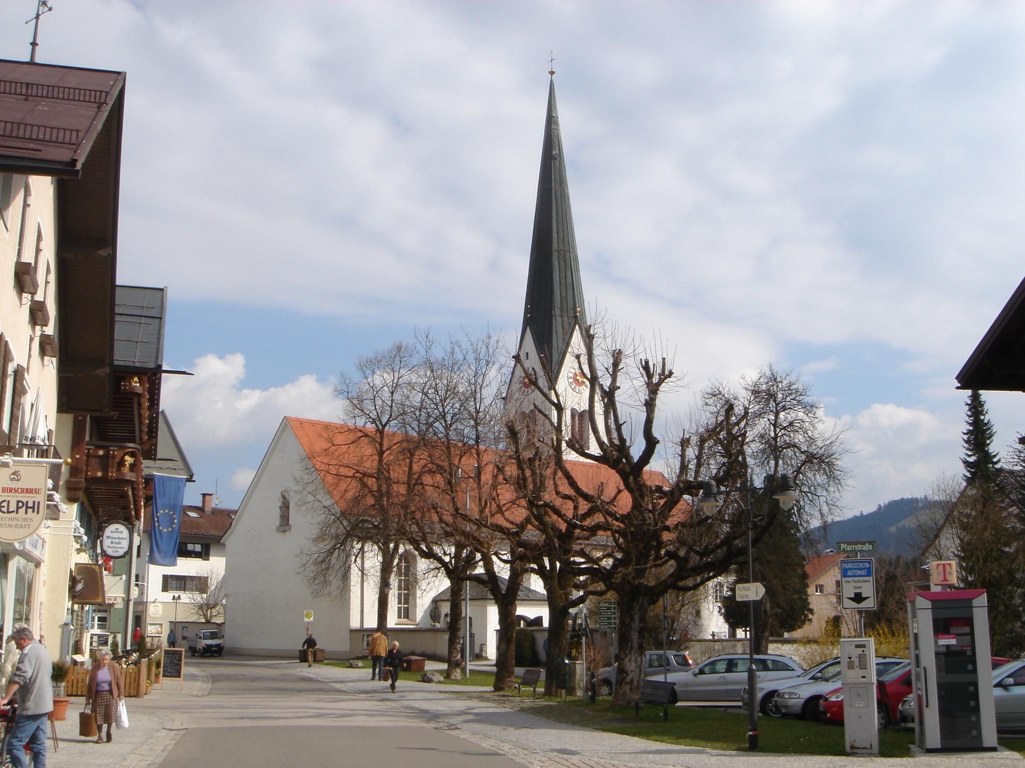Ein Bild der Kirche in Fischen mit spitzem Turm, im Vordergrund Bäume ohne Blätter.