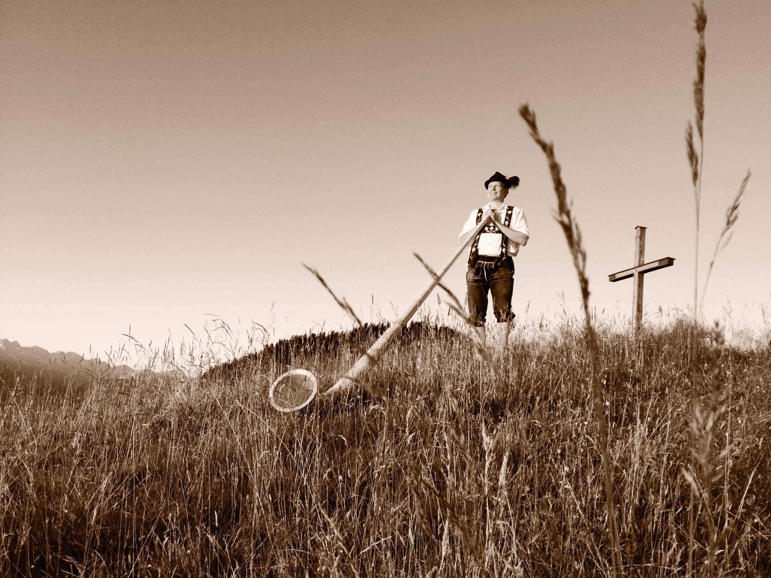 Eine sepiafarbene Aufnahme einer Person in traditioneller bayerischer Tracht, die auf einem grasbewachsenen Hügel steht und ein Alphorn bläst.