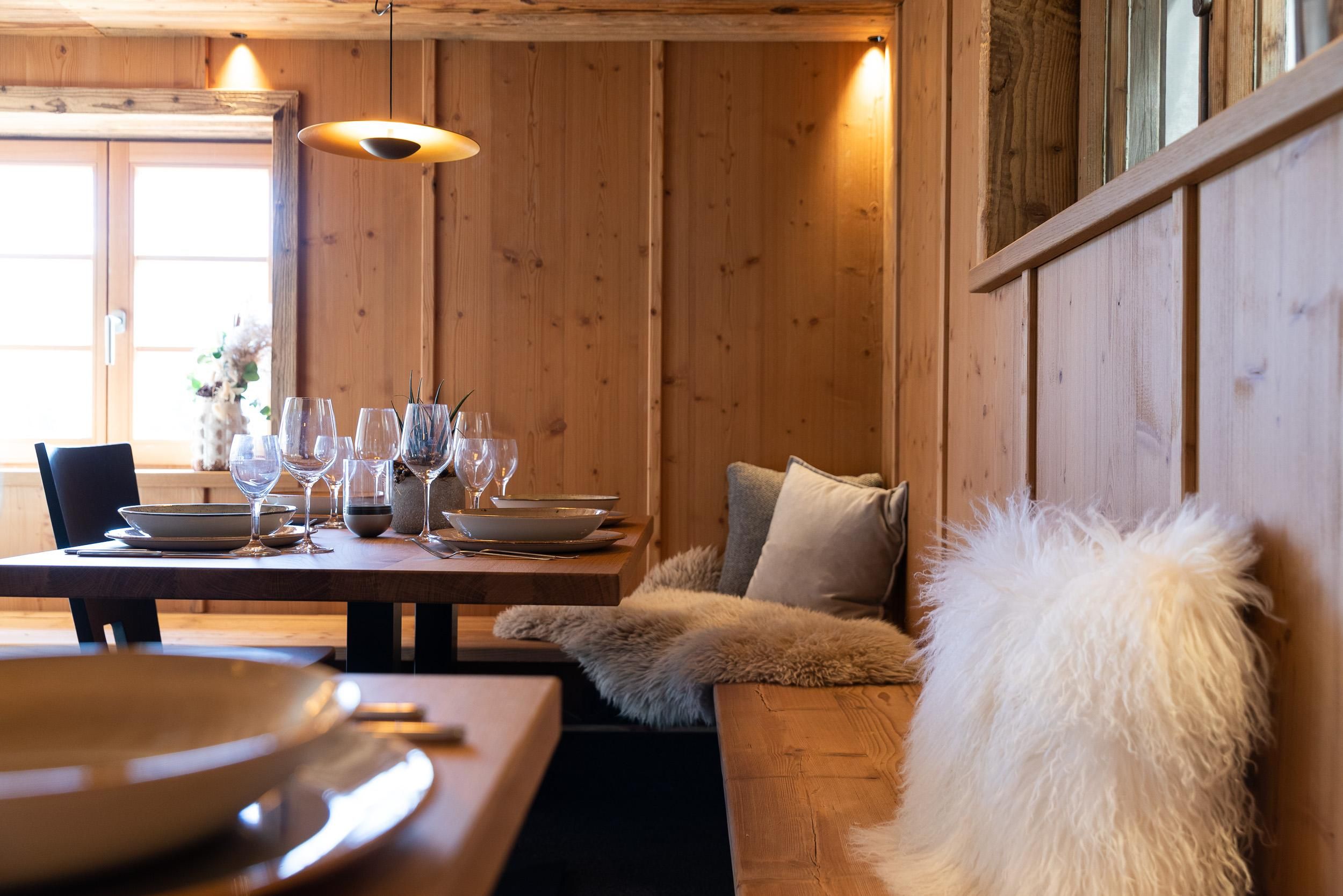Gemütlicher Essbereich mit Holztisch und eingedecktem Platz. Bank mit hellem Schaffell und Kissen.