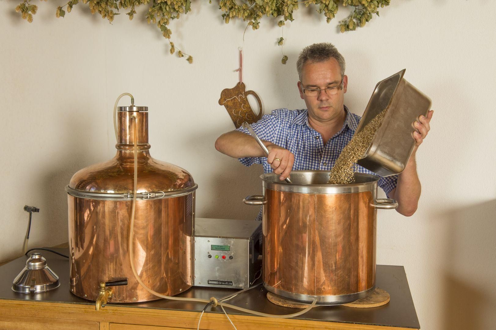 Mann schüttet Malz in Kupferkessel beim Bierbrauen. Zweiter Kessel, Steuerung, Hopfen, Krug.