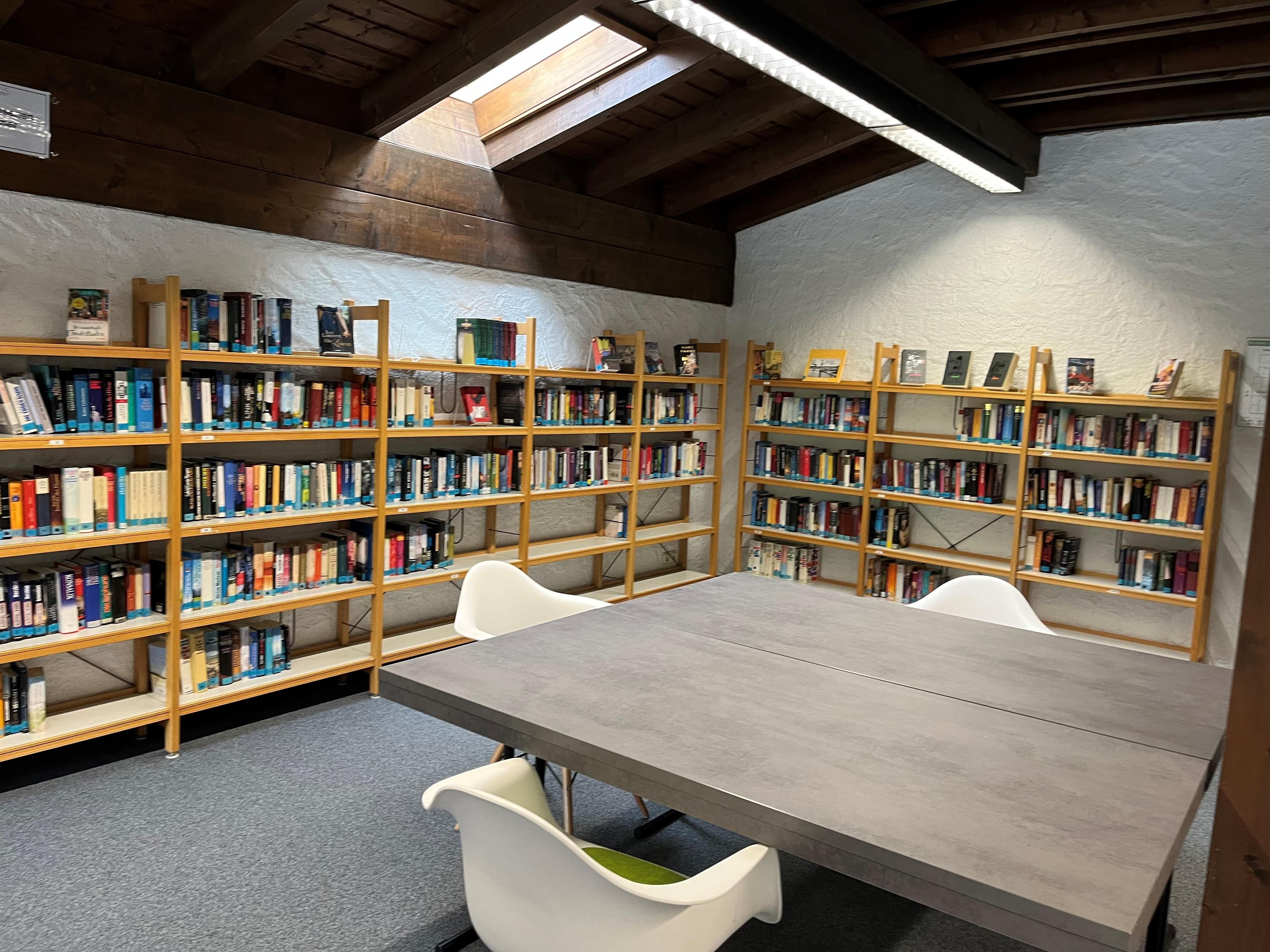 Innenraum der Gemeindebücherei Obermaiselstein. Holzregale gefüllt mit zahlreichen Büchern. Ein großer, grauer Tisch mit weißen Stühlen in der Mitte des Raumes.