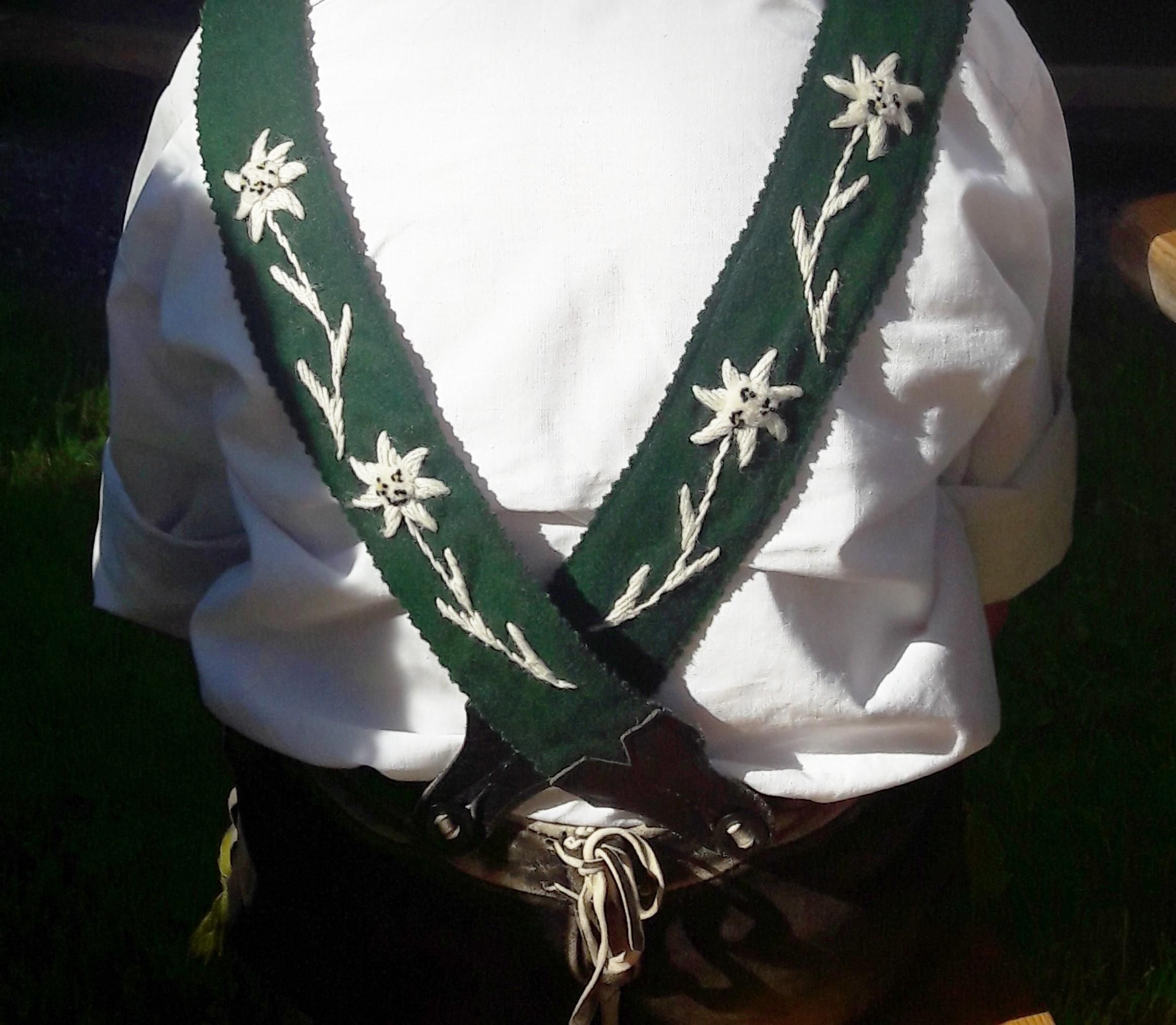 Detailaufnahme der Rückansicht einer Person, die eine weiße Trachtenbluse und grüne Lederhosen-Hosenträger mit weißen Edelweiß-Stickereien trägt.