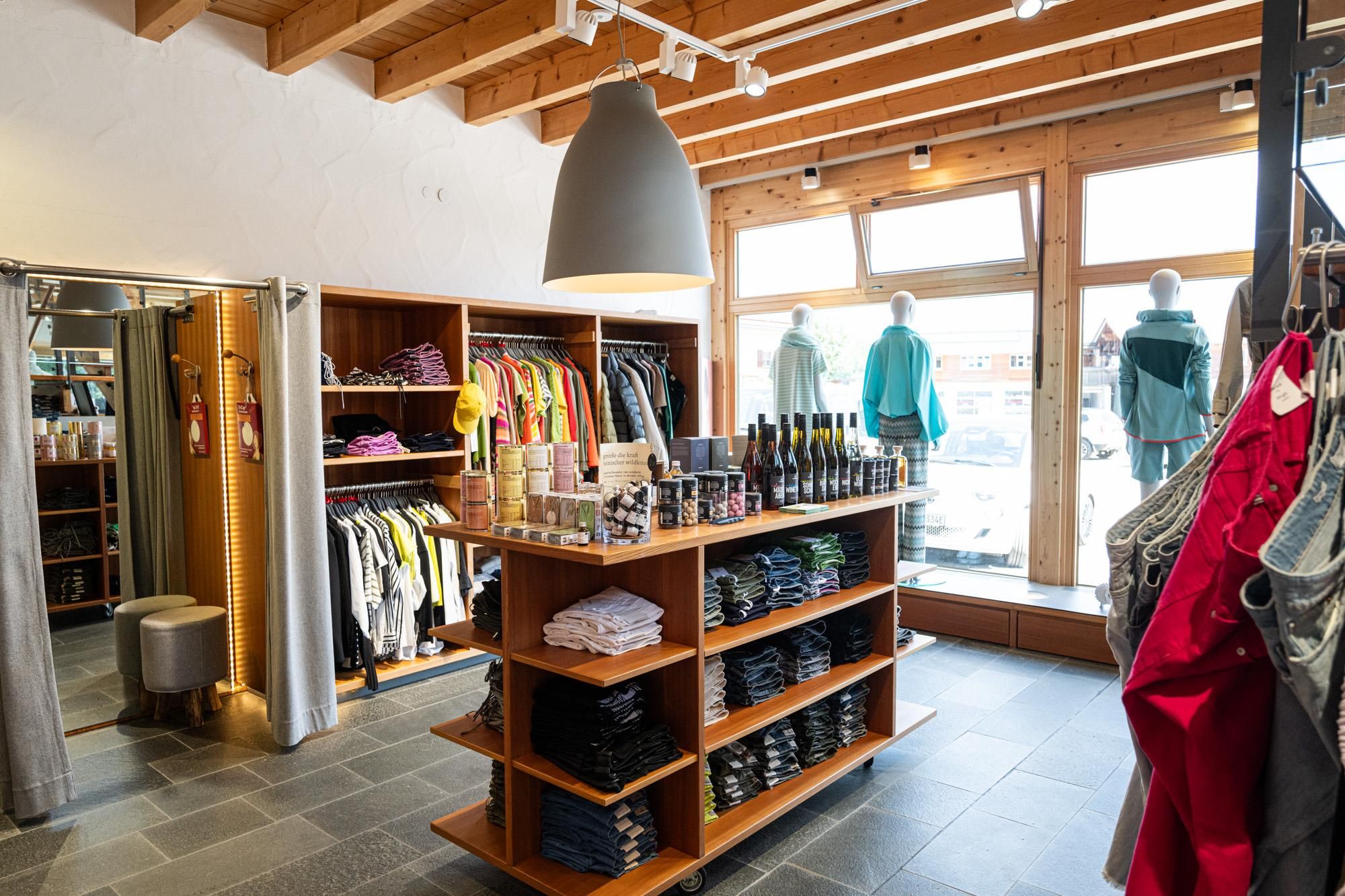 Holzregale mit farbenfroher Kleidung, gestapelten Textilien und Accessoires. Schaufenster mit Mannequins in modischer Bekleidung.