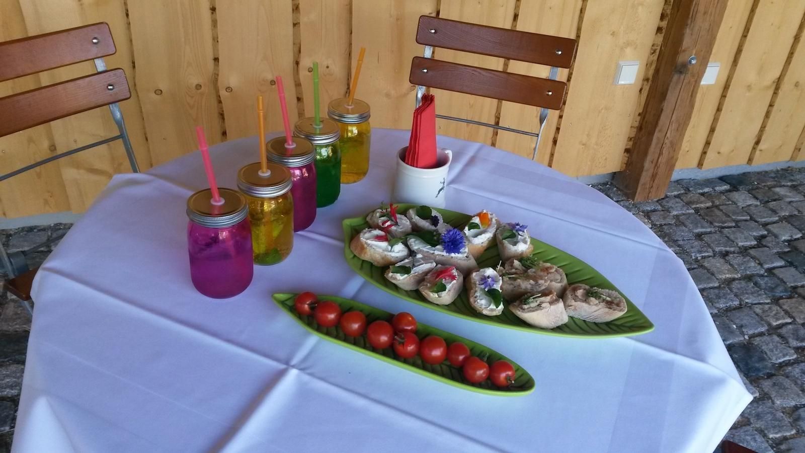Tisch mit weißer Decke, belegten Broten, Tomaten und bunten Getränken. Zwei Holzstühle stehen am Tisch.