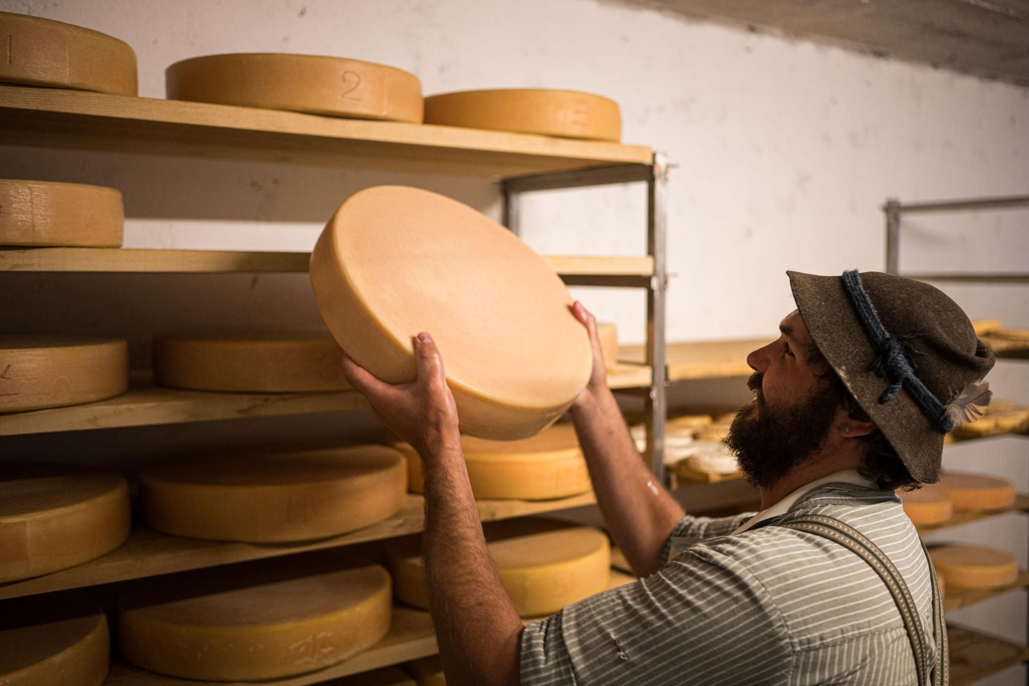 Ein Senner wendet einen Käselaib. In einem Regal reifen weitere Käselaibe.