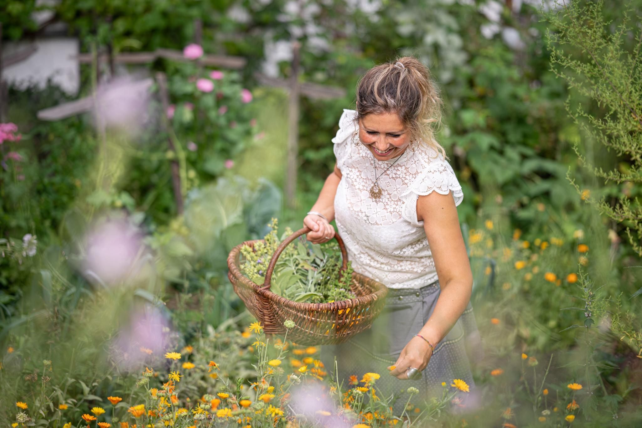 Eine lächelnde Frau mit hochgesteckten blonden Haaren trägt ein weißes Oberteil und einen grauen Rock. Sie hält einen gefüllten Weidenkorb in einem blühenden Garten.