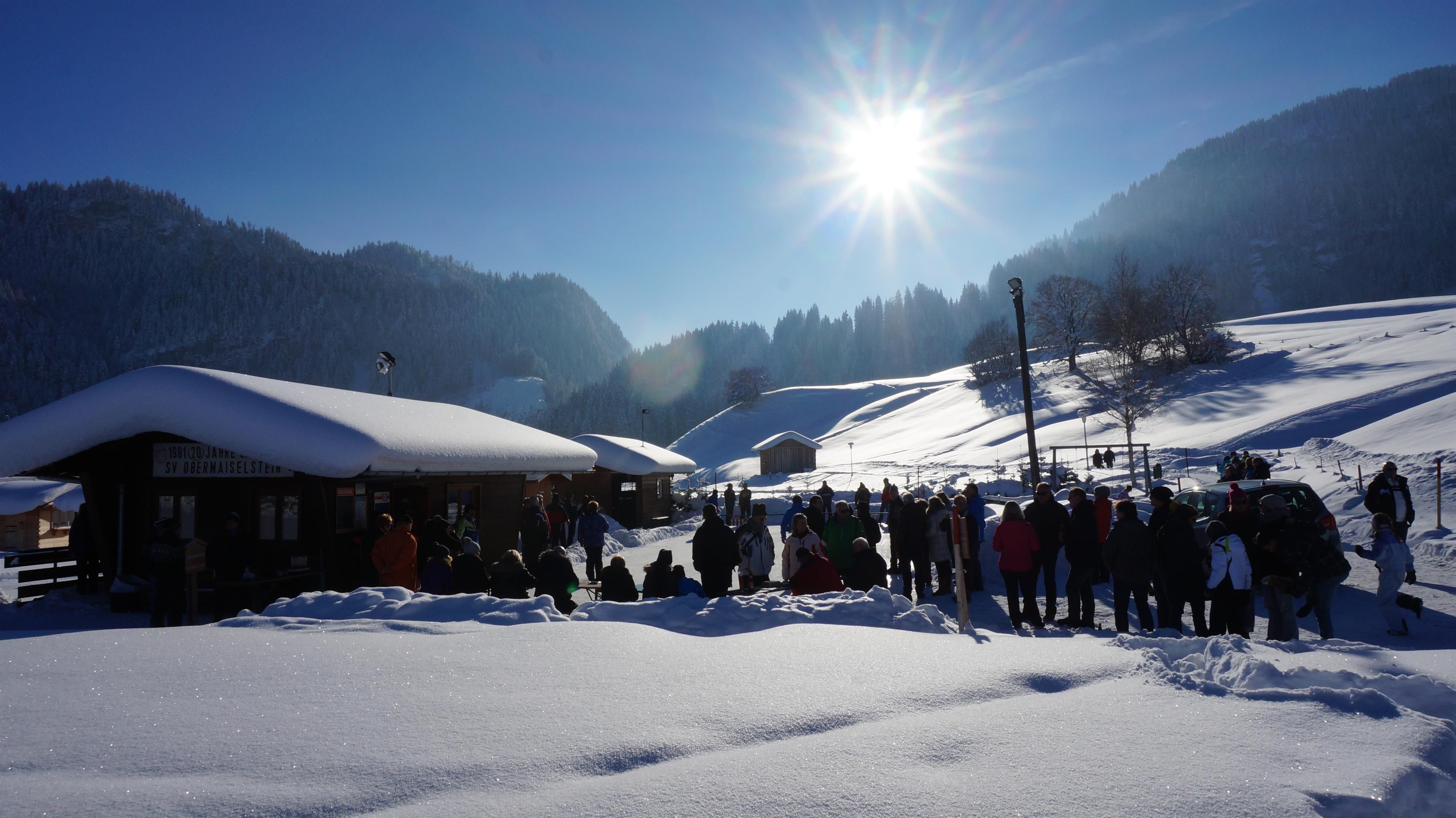 Sonniger Wintertag am Eisstockplatz. Verschneite Landschaft mit Holzhütten und einer Gruppe von Menschen.