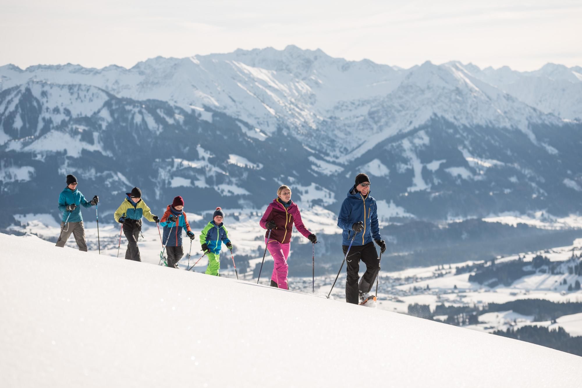 Sechs Personen bei einer Schneeschuhwanderung. Sie gehen in einer Reihe einen verschneiten Hang hinauf. Im Hintergrund verschneite Bergkulisse.