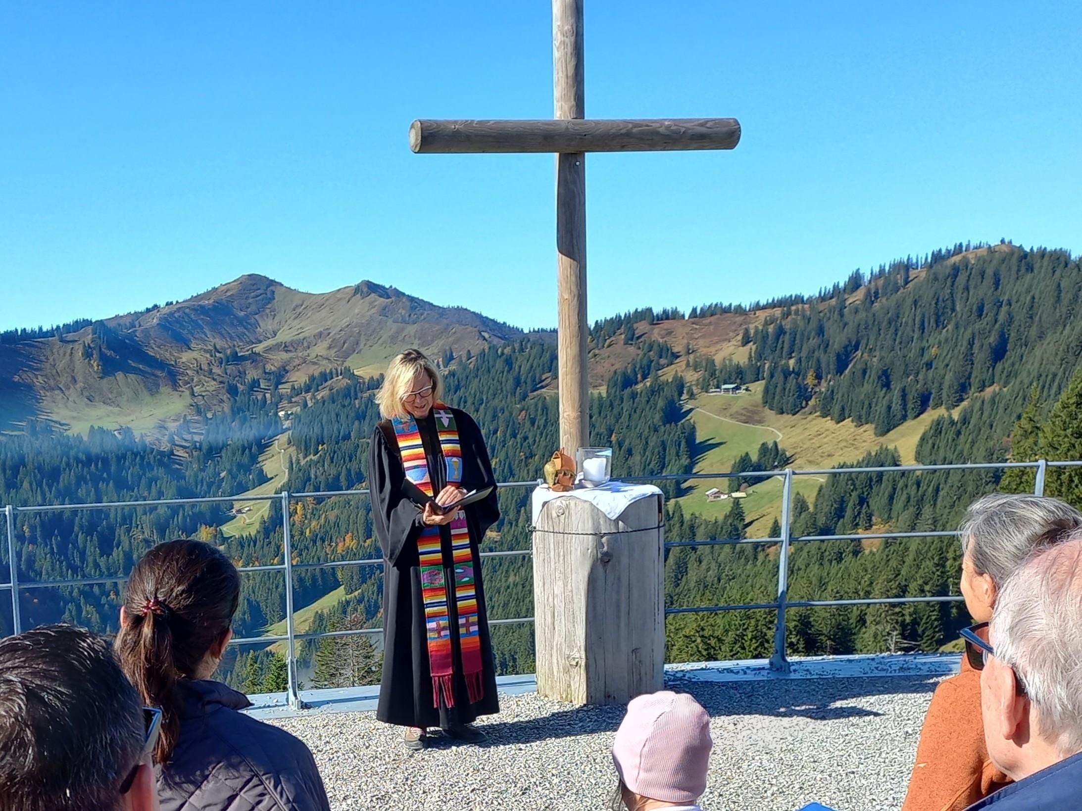 Eine Person in einer schwarzen Kleidung steht vor einem Kreuz in den Bergen. Im Hintergrund sind grüne Hügel und ein klarer blauer Himmel zu sehen.