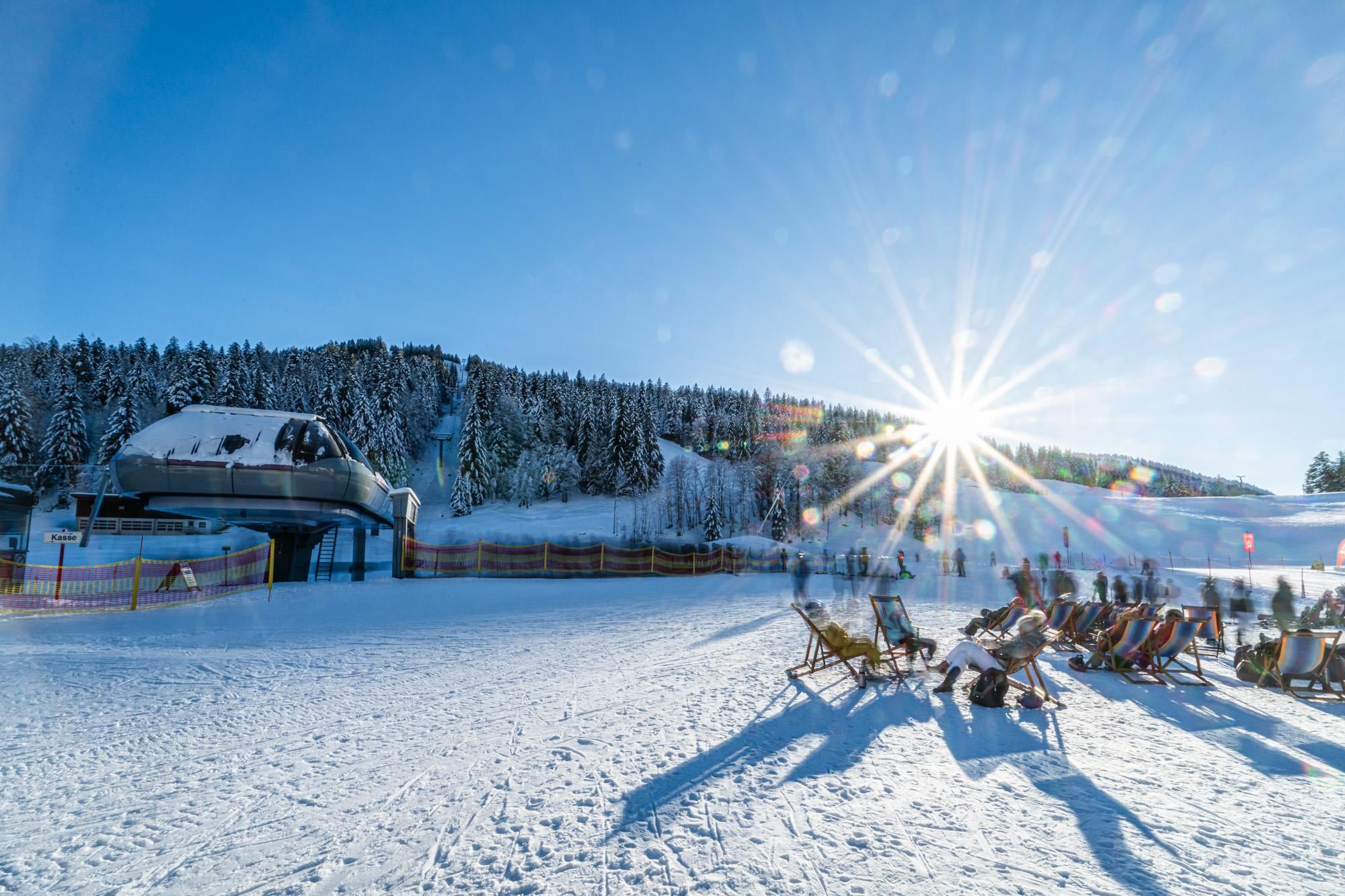 Wintersonne im Skigebiet Balderschwang: Im Vordergrund entspannen Skifahrer in Liegestühlen, im Hintergrund eine Sesselbahn und verschneite Bäume.