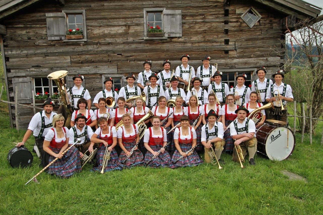 Eine Trachtenkapelle mit 45 Musikern und Instrumenten posiert vor alpiner Berglandschaft auf einer grünen Wiese.