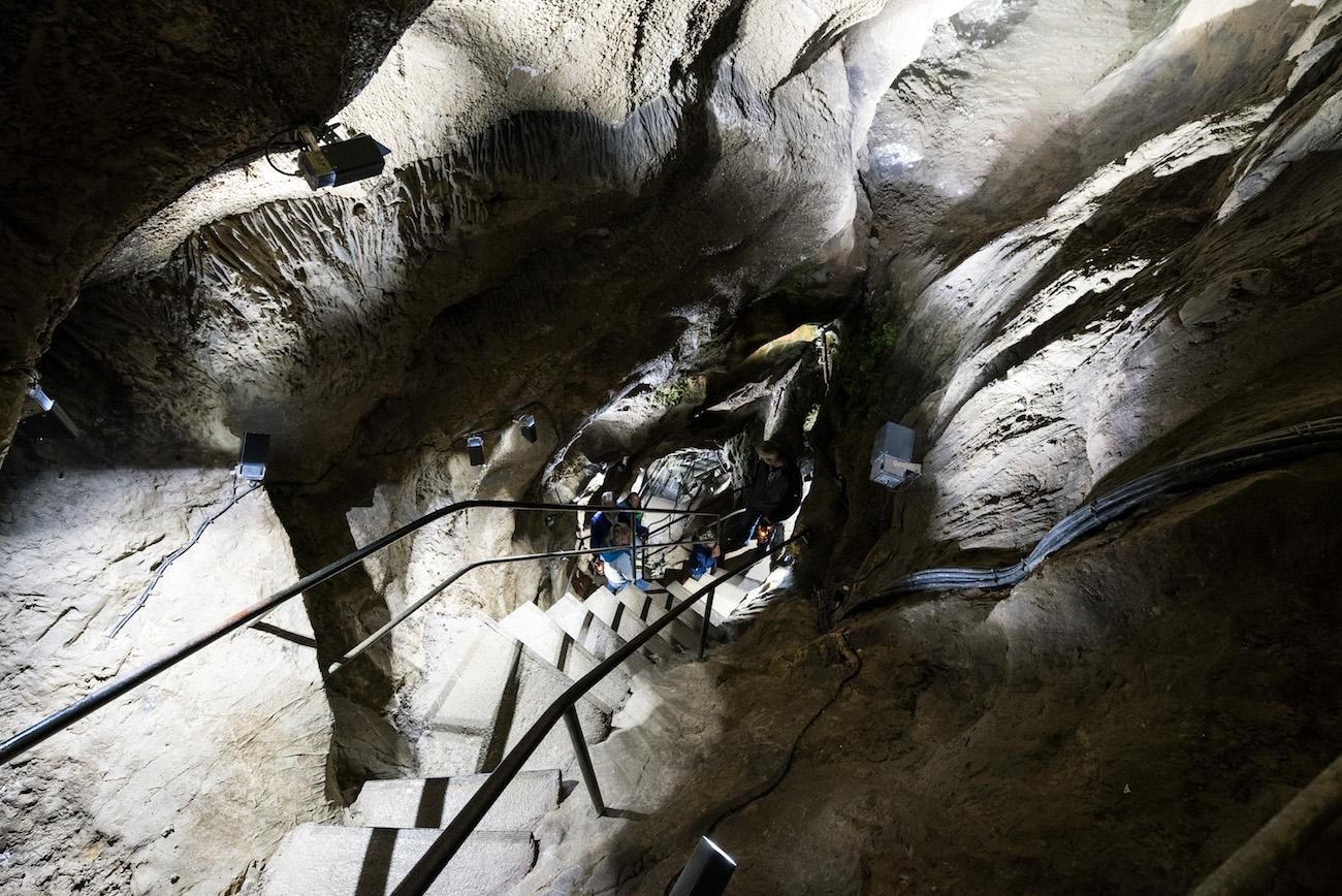 Blick in die beleuchtete Sturmannshöhle. Steile Treppe mit Geländer führt nach unten. Rauhe Felswände umgeben den Weg. Menschen sind in der Tiefe erkennbar.