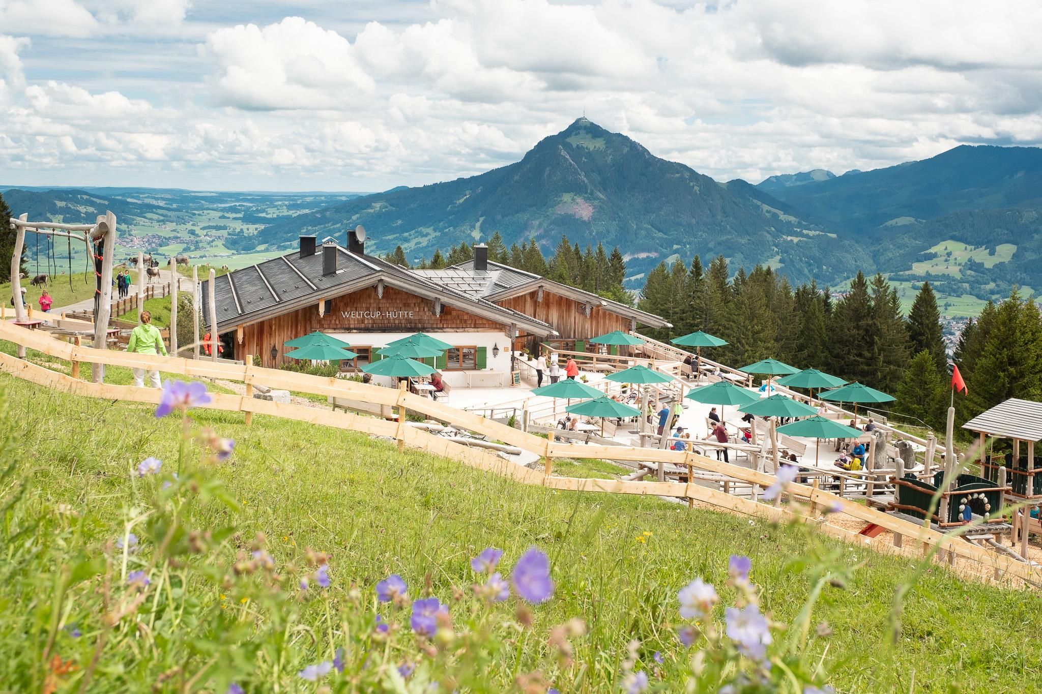<p>Bergrestaurant Weltcup-Hütte Ofterschwang mit Holzbalkon, grünen Sonnenschirmen und Gästen. Im Vordergrund blühende Wiese, im Hintergrund Bergpanorama unter bewölktem Himmel.</p>