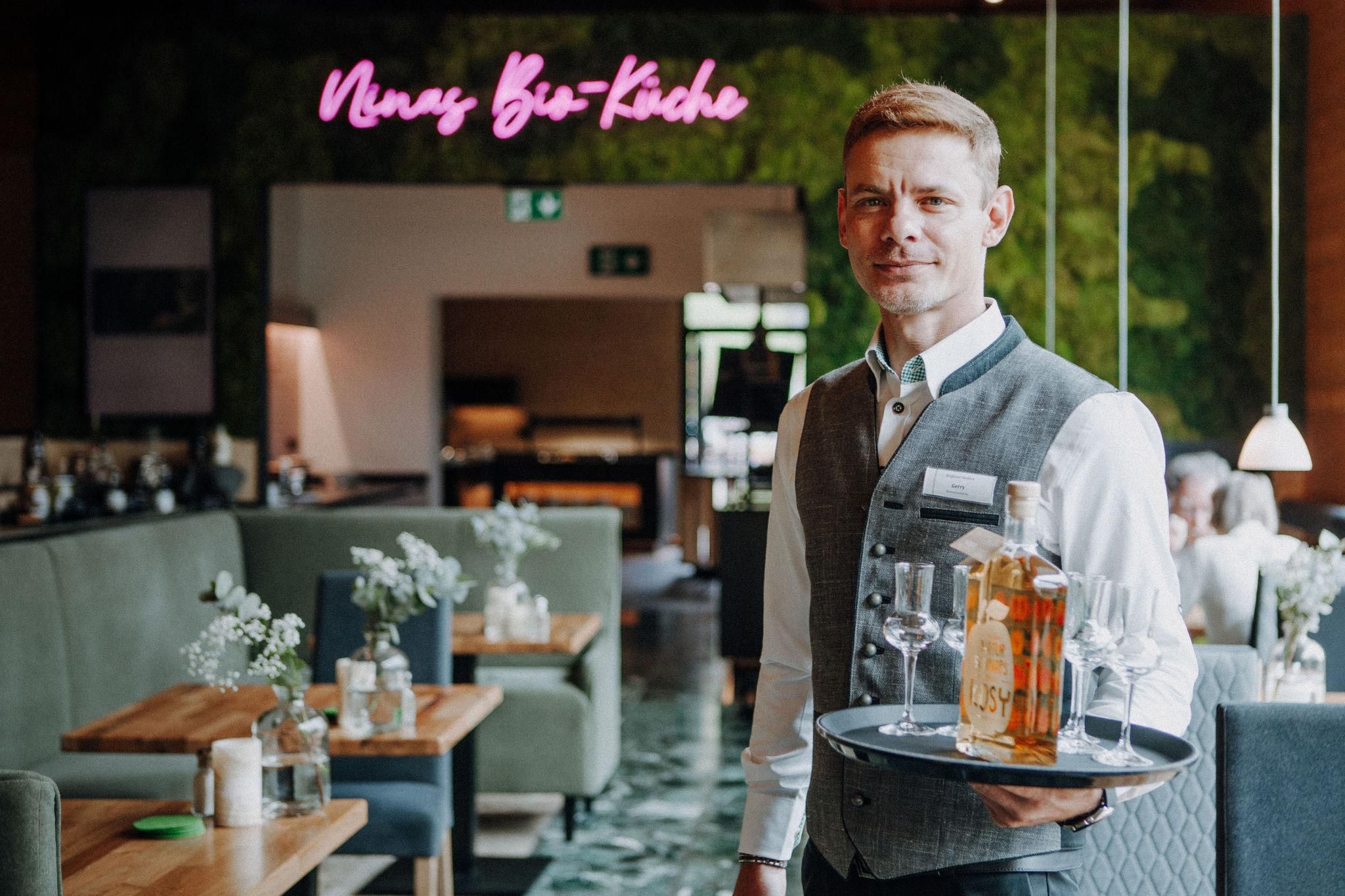 Ein Kellner steht in einem modern eingerichteten Raum und hält ein Tablett mit Gläsern und einer Flasche Schnaps in der Hand.