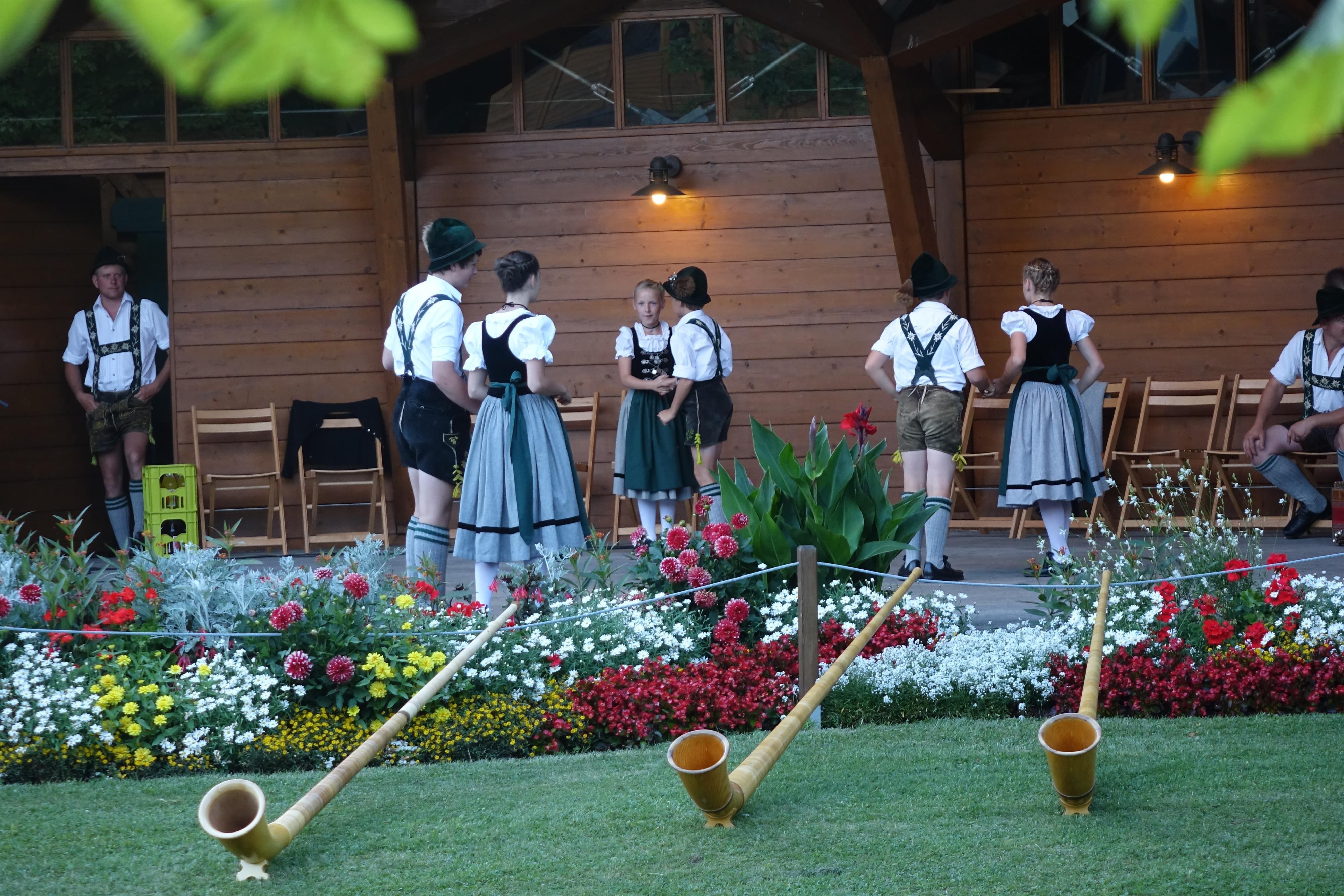 Die Schuhplattlergruppe in Tracht bei einem Auftritt im Kurpark auf der Bühne. Im Vordergrund liegen Alphörner im Gras.