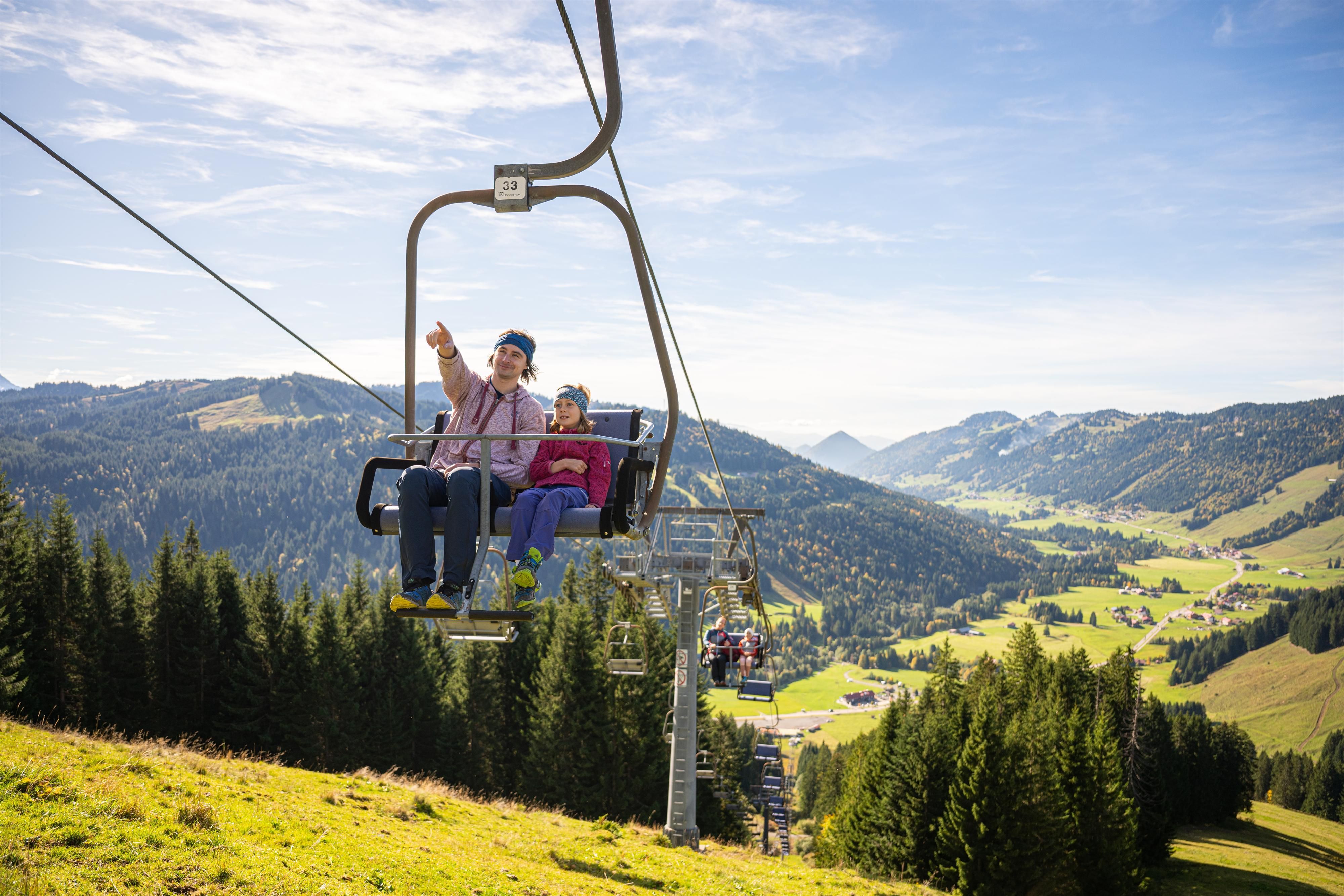Ein Erwachsener und ein Kind sitzen gemeinsam auf einem Sessellift und fahren über eine grüne, hügelige Berglandschaft mit Wäldern und verstreuten Häusern. Der Erwachsene zeigt in die Ferne, während beide entspannt wirken und unter einem hellen, leicht bewölkten Himmel die Aussicht genießen.