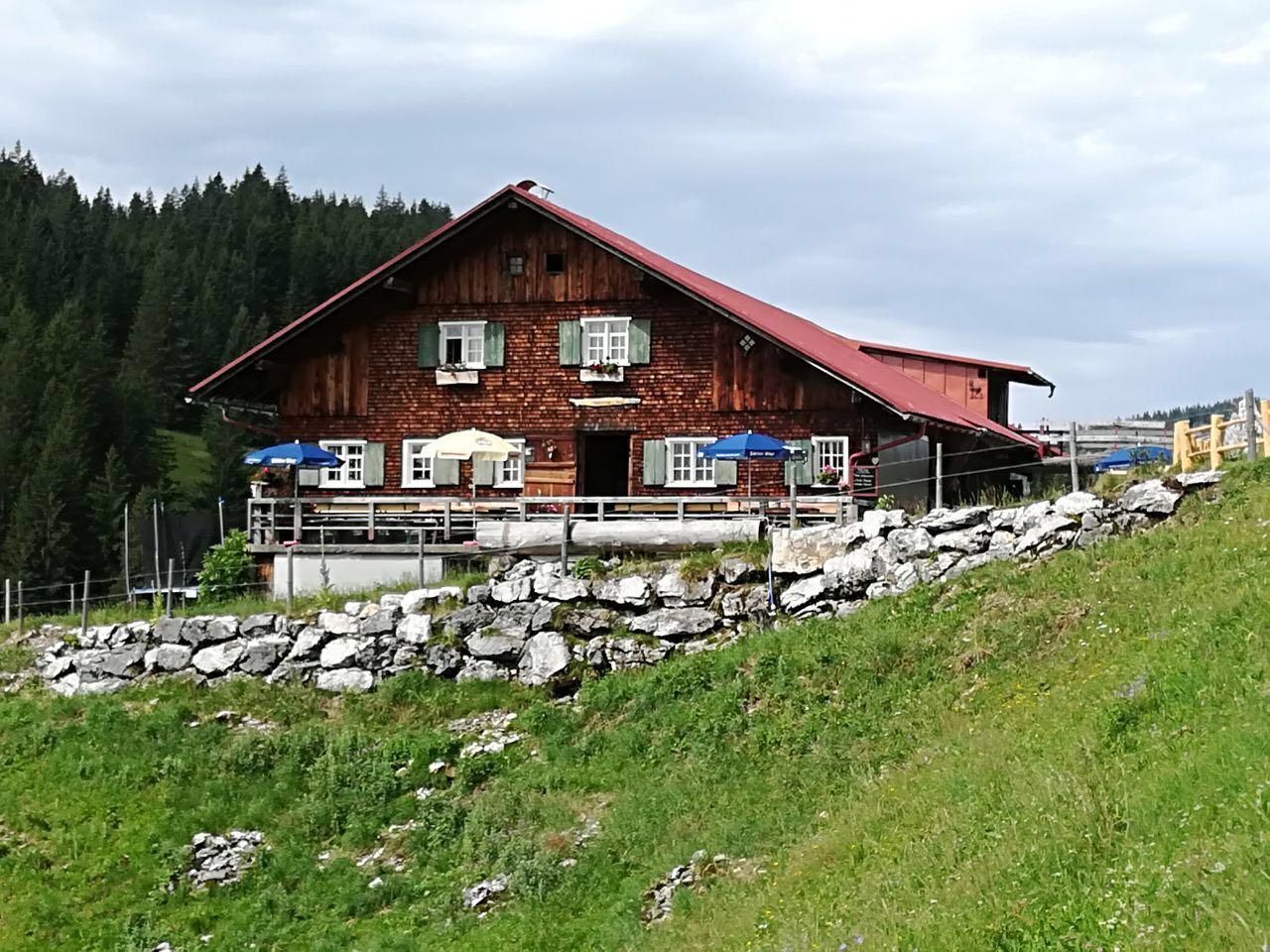 Braunes Holzhaus mit rotem Dach, grünen Fensterläden und blauen Sonnenschirmen auf einer Terrasse mit Steinstützmauer in grüner Landschaft.