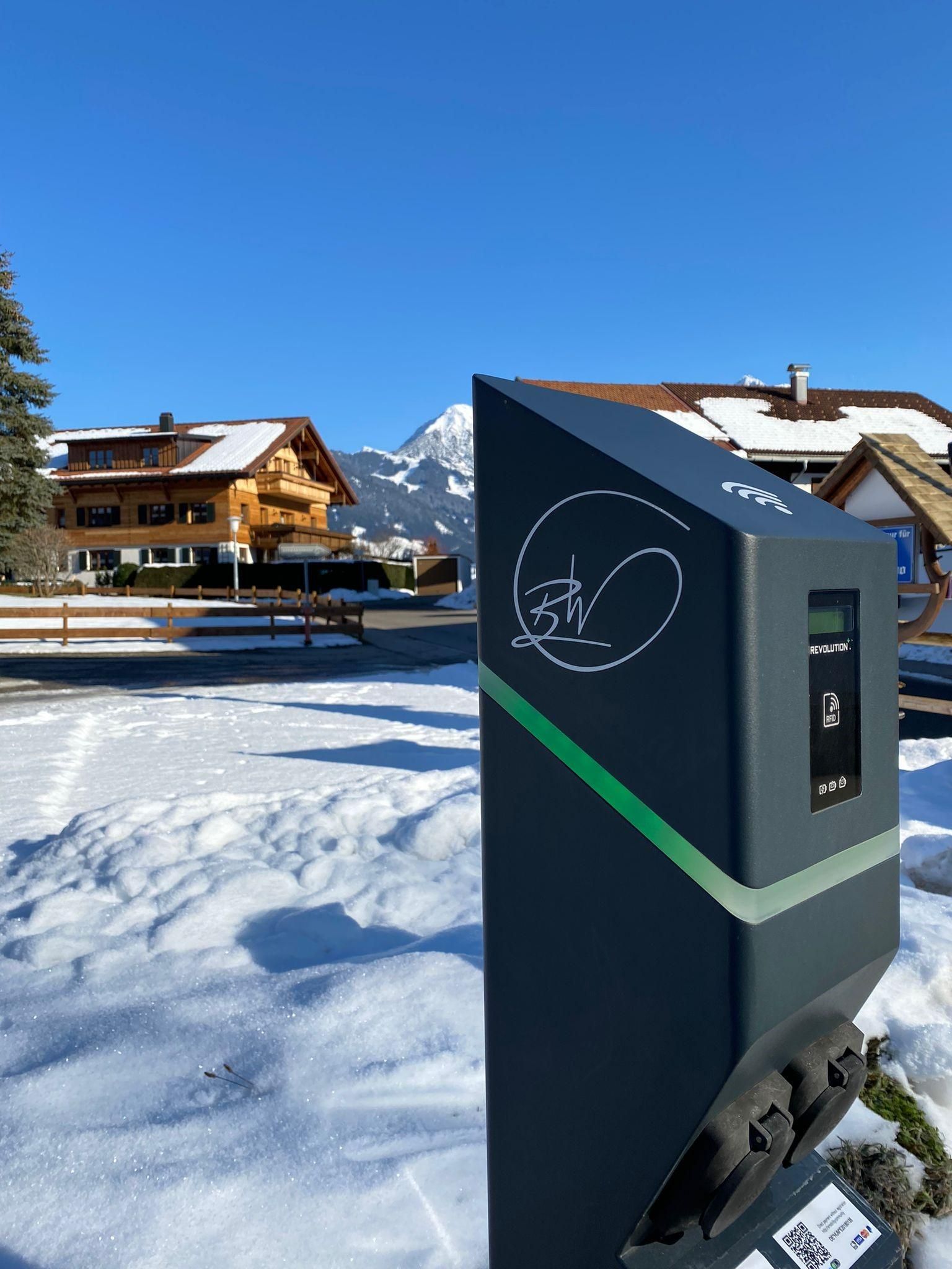 Eine moderne, dunkelgraue E-Auto-Ladestation mit grünen Akzenten im Schnee. Im Hintergrund sind verschneite Gebäude und ein klarer, blauer Himmel zu sehen.