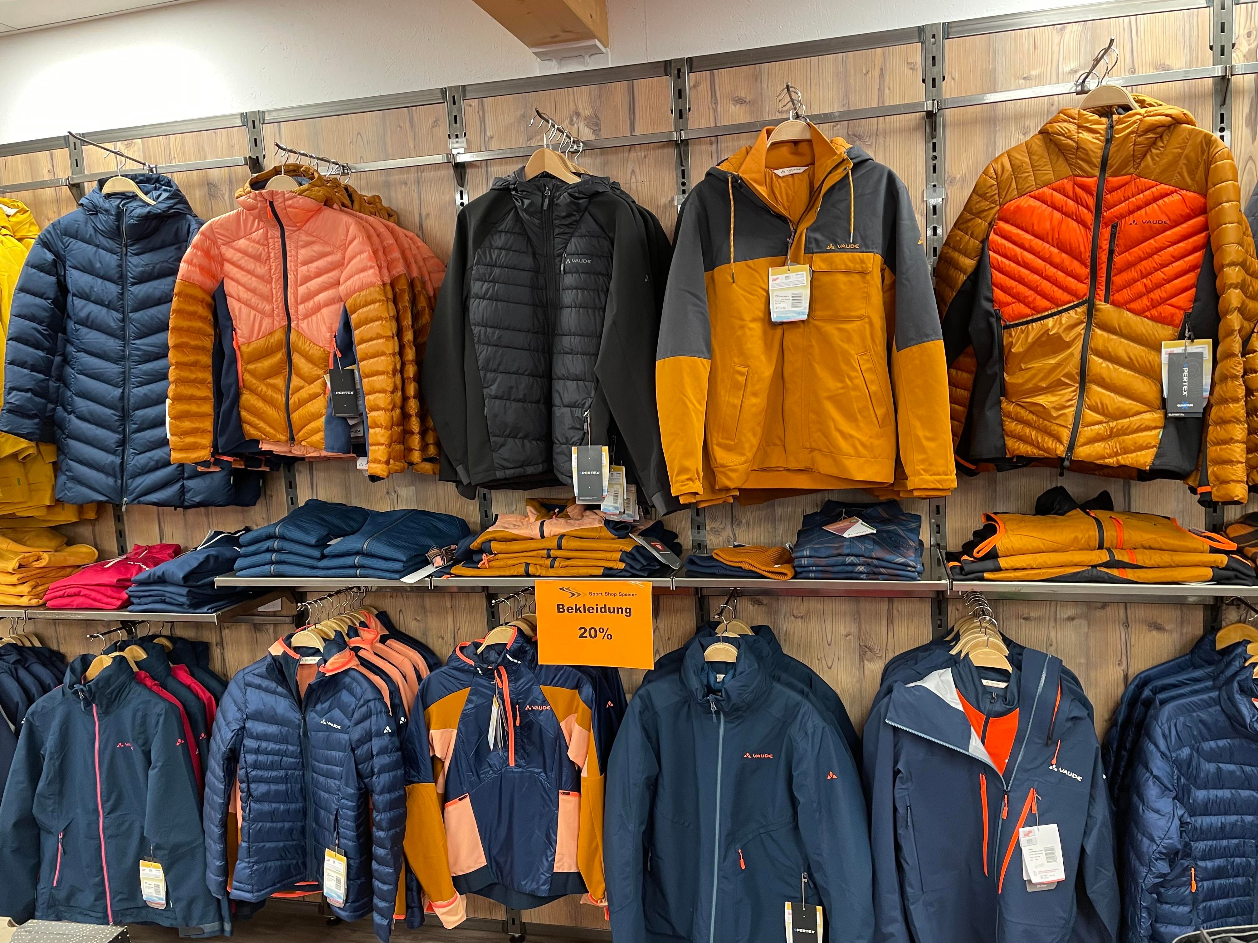 Bekleidungsgeschäft, zwei Reihen Jacken an Kleiderbügeln, verschiedene Farben und Stile. Untere Reihe gefaltete Kleidung. Schild "Bekleidung 20%". Helle Holzwand im Hintergrund.