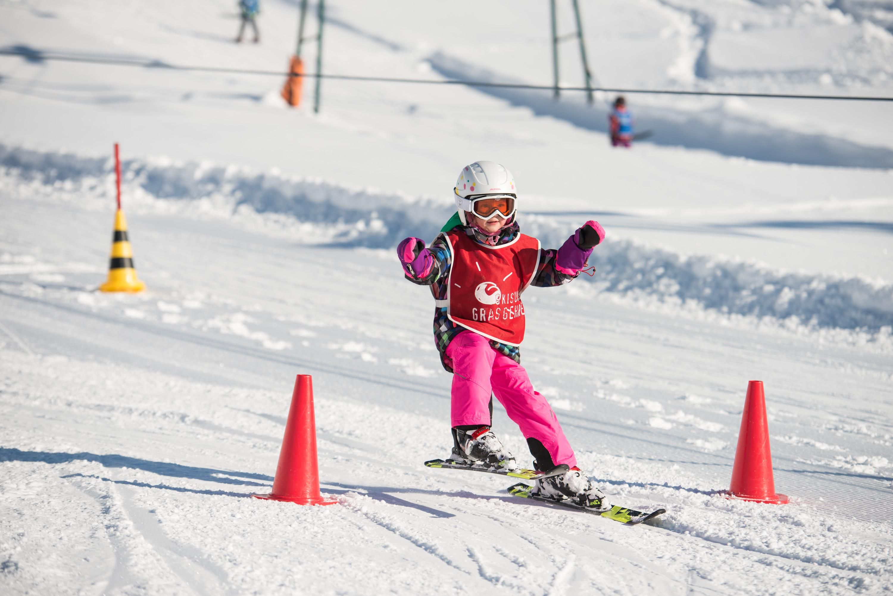 Ein Kind in Skikleidung, Helm und Skibrille fährt Ski auf einer verschneiten Piste zwischen zwei roten Kegeln.
