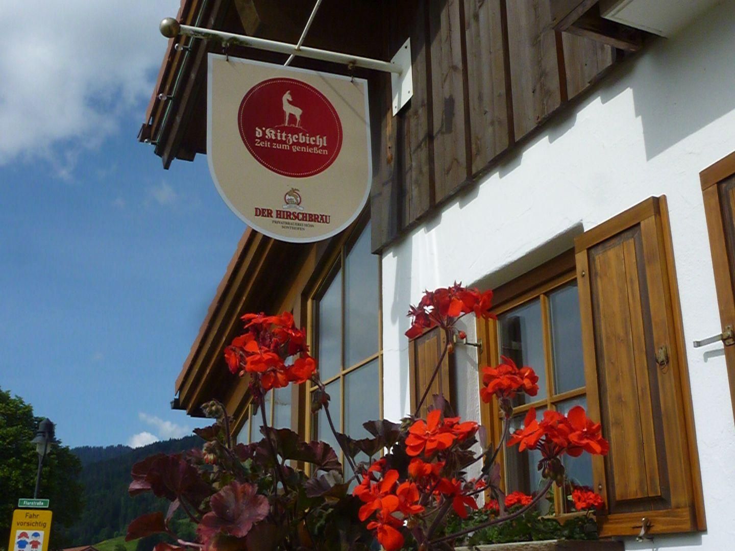 Schild "D'Kitzebichl - Zeit zum Genießen" mit Hirschlogo am Restaurant Kitzbichl in Obermaiselstein. Weißes Gebäude mit braunen Fensterläden und roten Geranien.